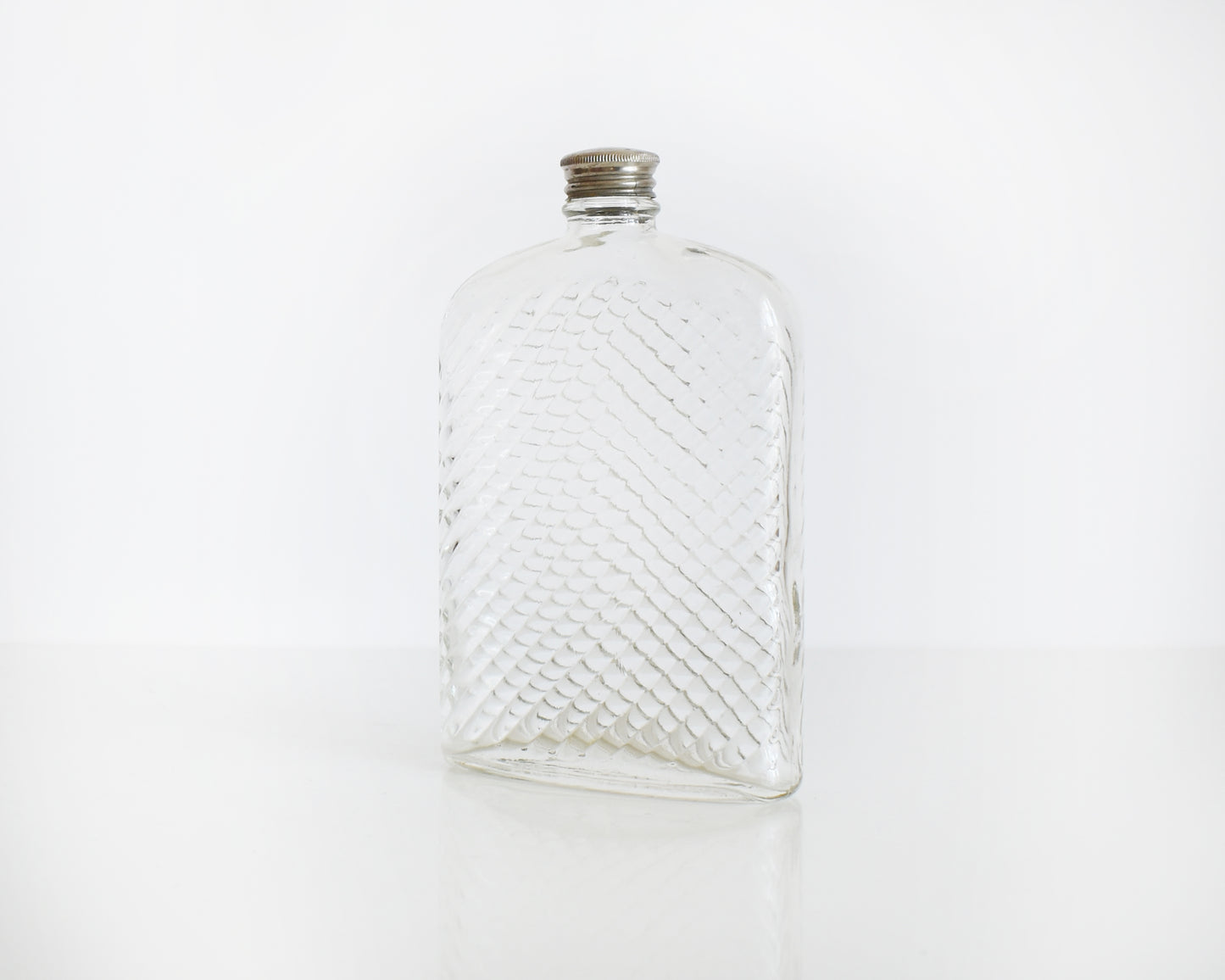 Side front view of a vintage glass flask that has a cut diamond pattern and a silver screw top. The flask is on a white table and a white background.