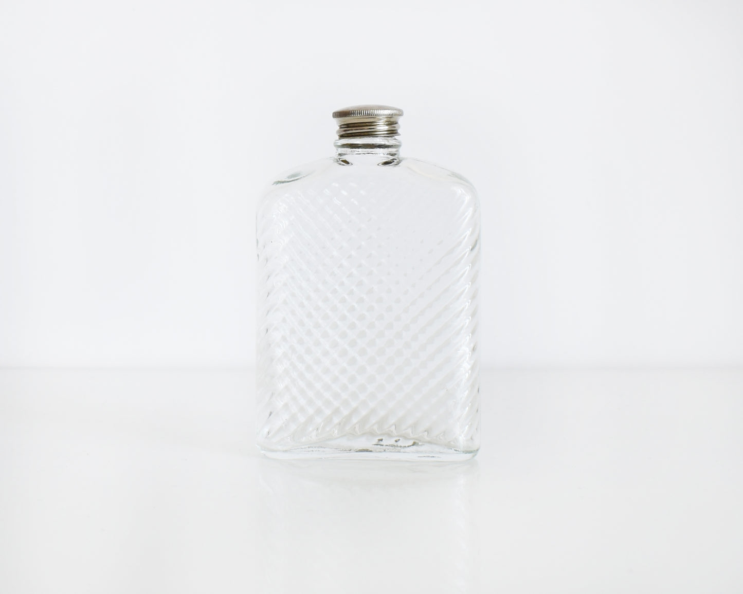Back view of a vintage glass flask that has a cut diamond pattern and a silver screw top. The flask is on a white table and a white background.