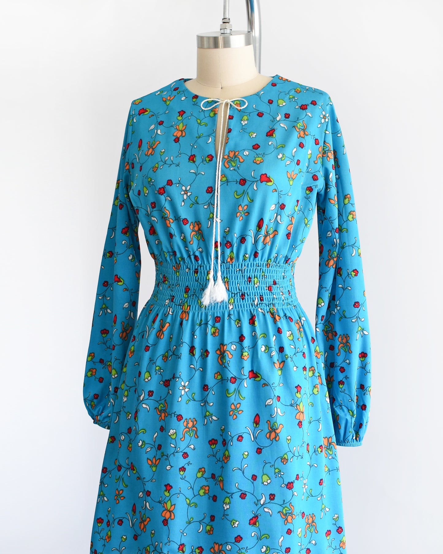 Side front view of a vintage blue dress with a vine floral print with tassel neckline, smocked waist, long sleeves, and a skirt that hits above the knees. The dress is modeled on a dress form.