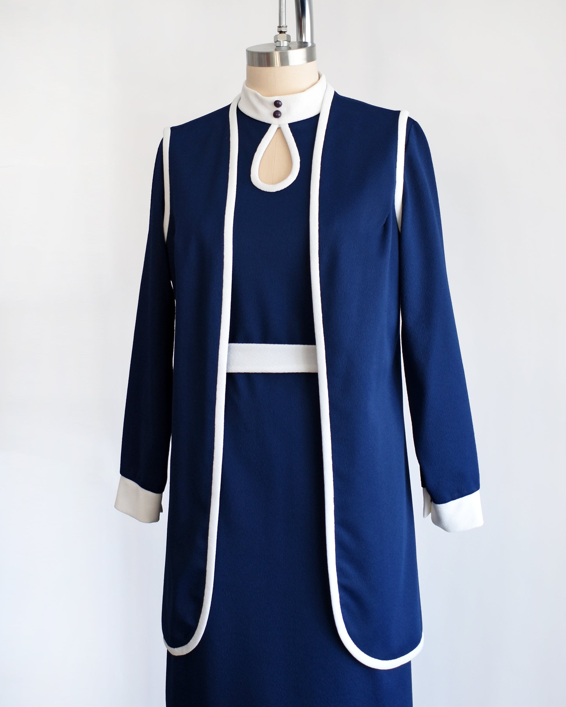 Side front view of a late 1960s to early 1970s mod two piece set features a navy blue long sleeve dress and matching vest and white belt.