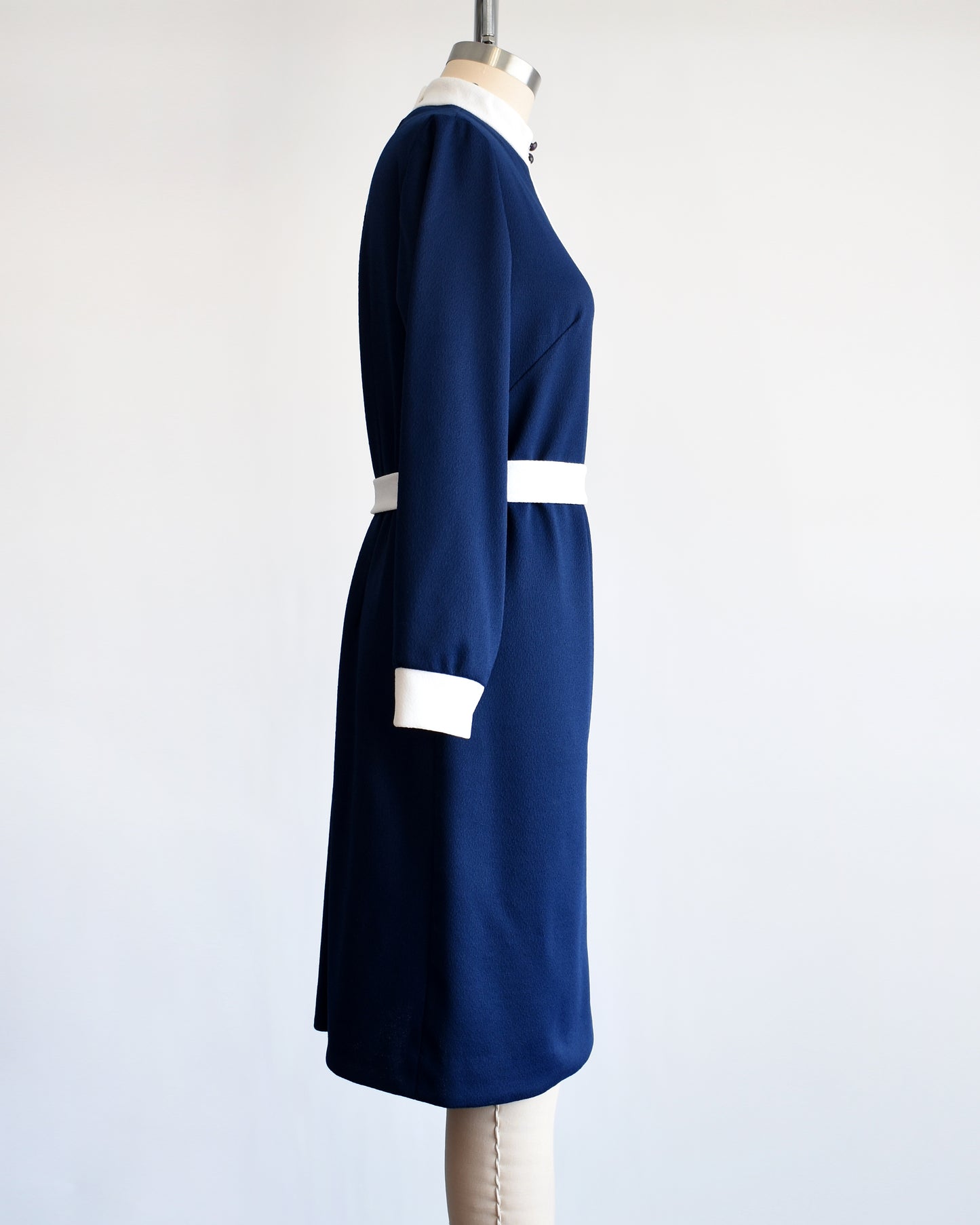 Side view of a late 1960s to early 1970s mod two piece set features a navy blue long sleeve dress and matching vest and white belt.