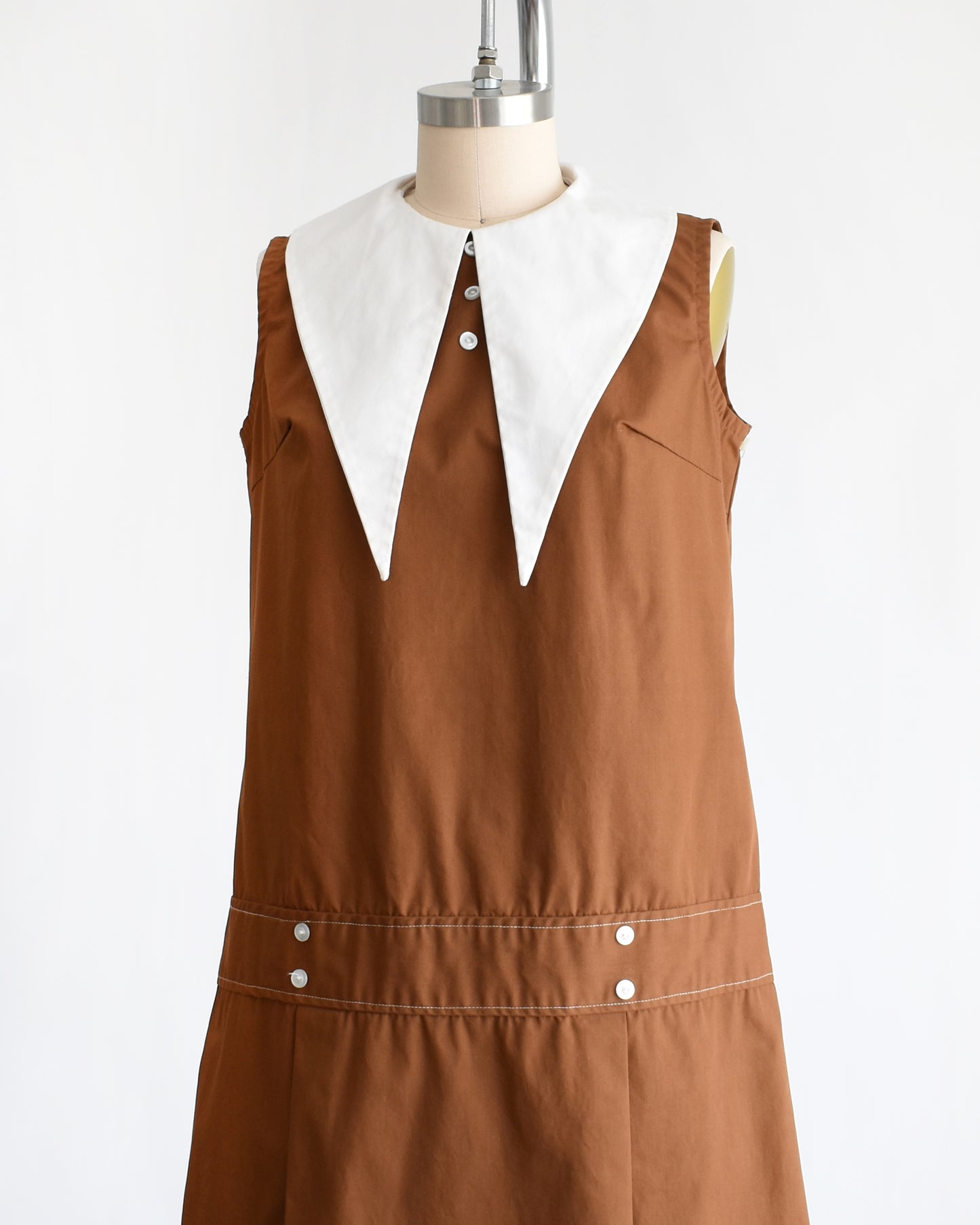 side front view of a vintage 1960s brown drop waist dress that has a large white dagger collar and decorative buttons on the front