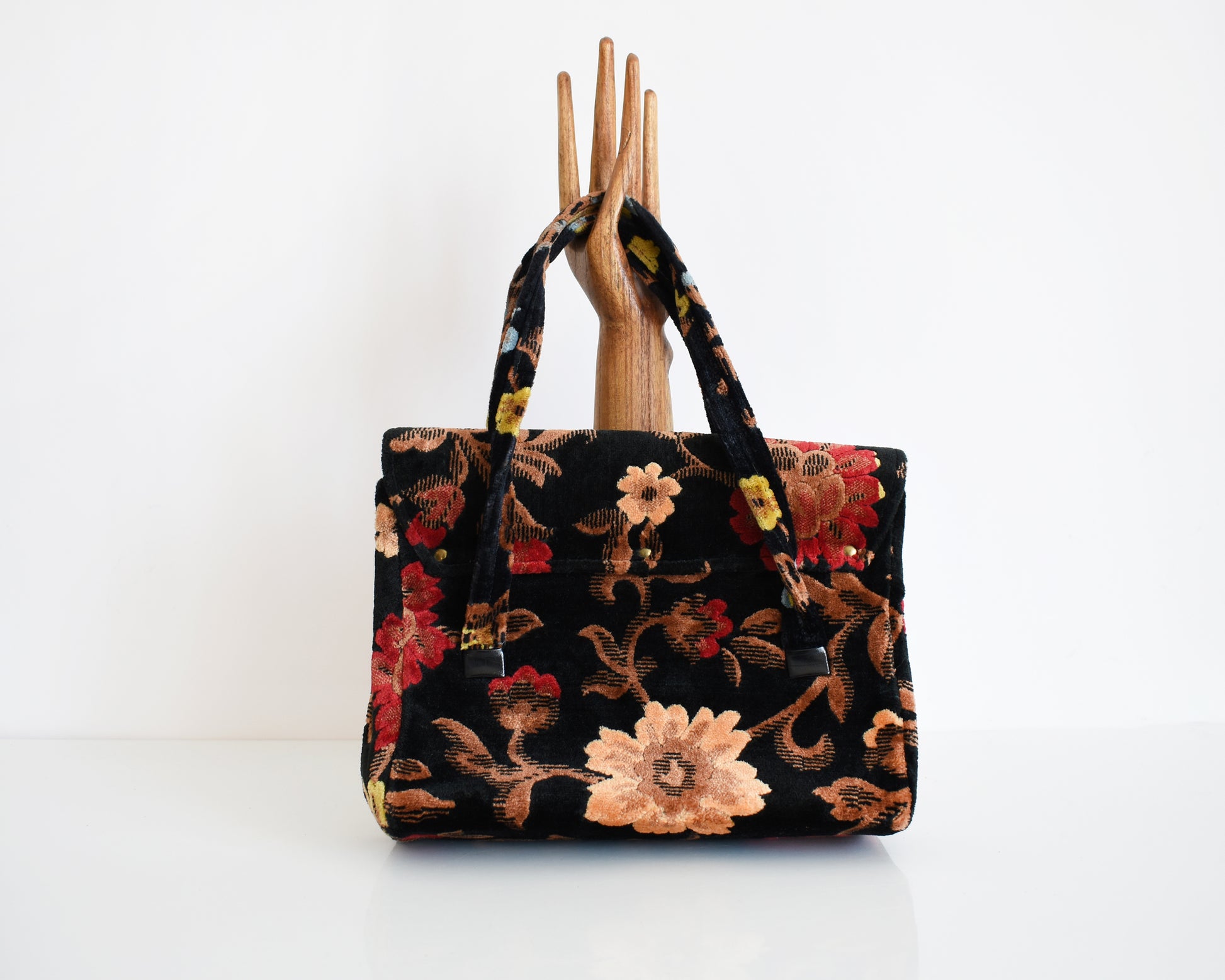 Back view of a vintage black floral velvet chenille handbag with gold tone hardware modeled with a wooden hand on a white table. The flowers are red, blue, yellow and have brown stems and leaves.