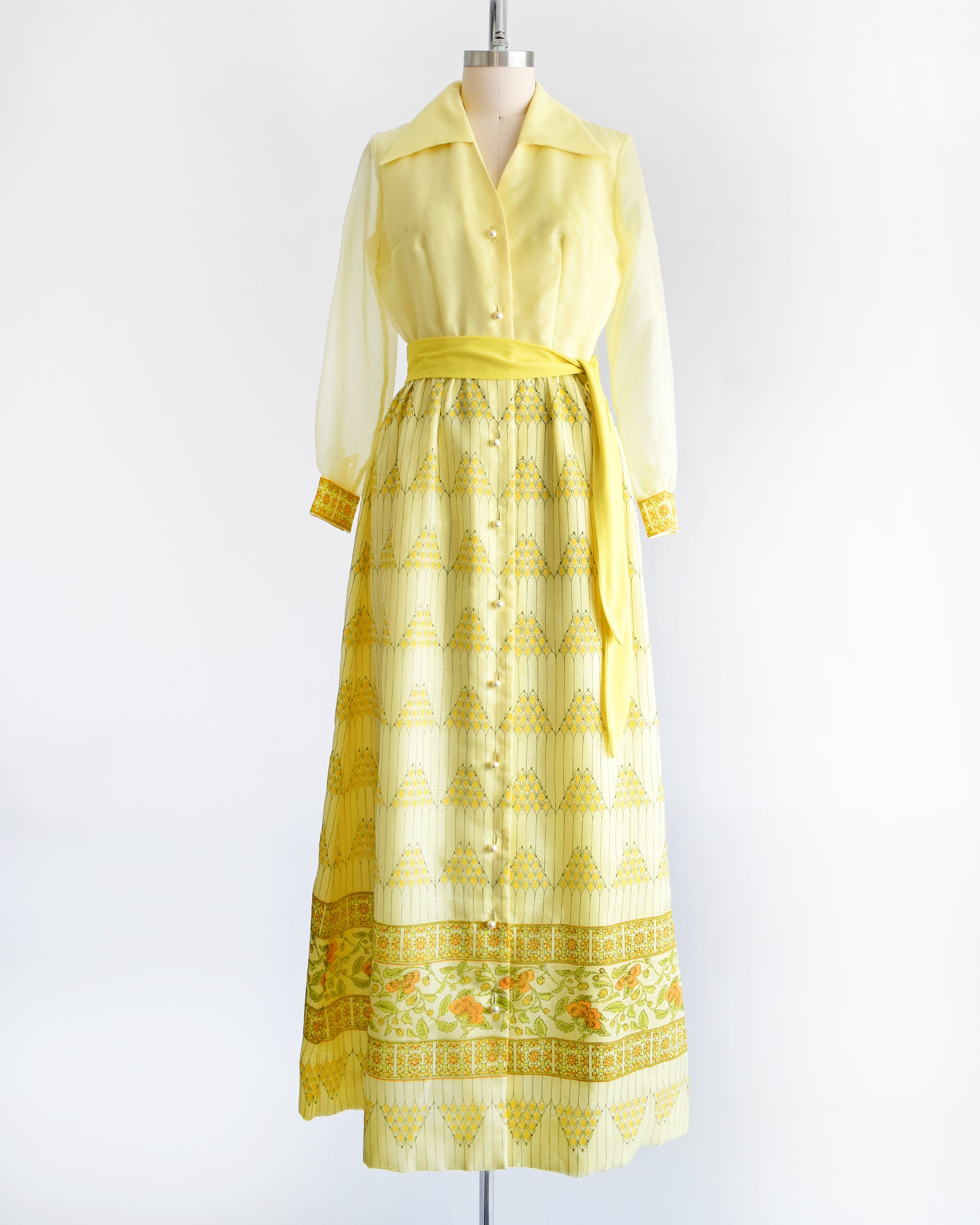 A yellow vintage maxi dress on a dress form. The dress features a collared neckline, semi sheer long sleeves, button down front, yellow sash belt, and a maxi skirt that has a pyramid and floral print.