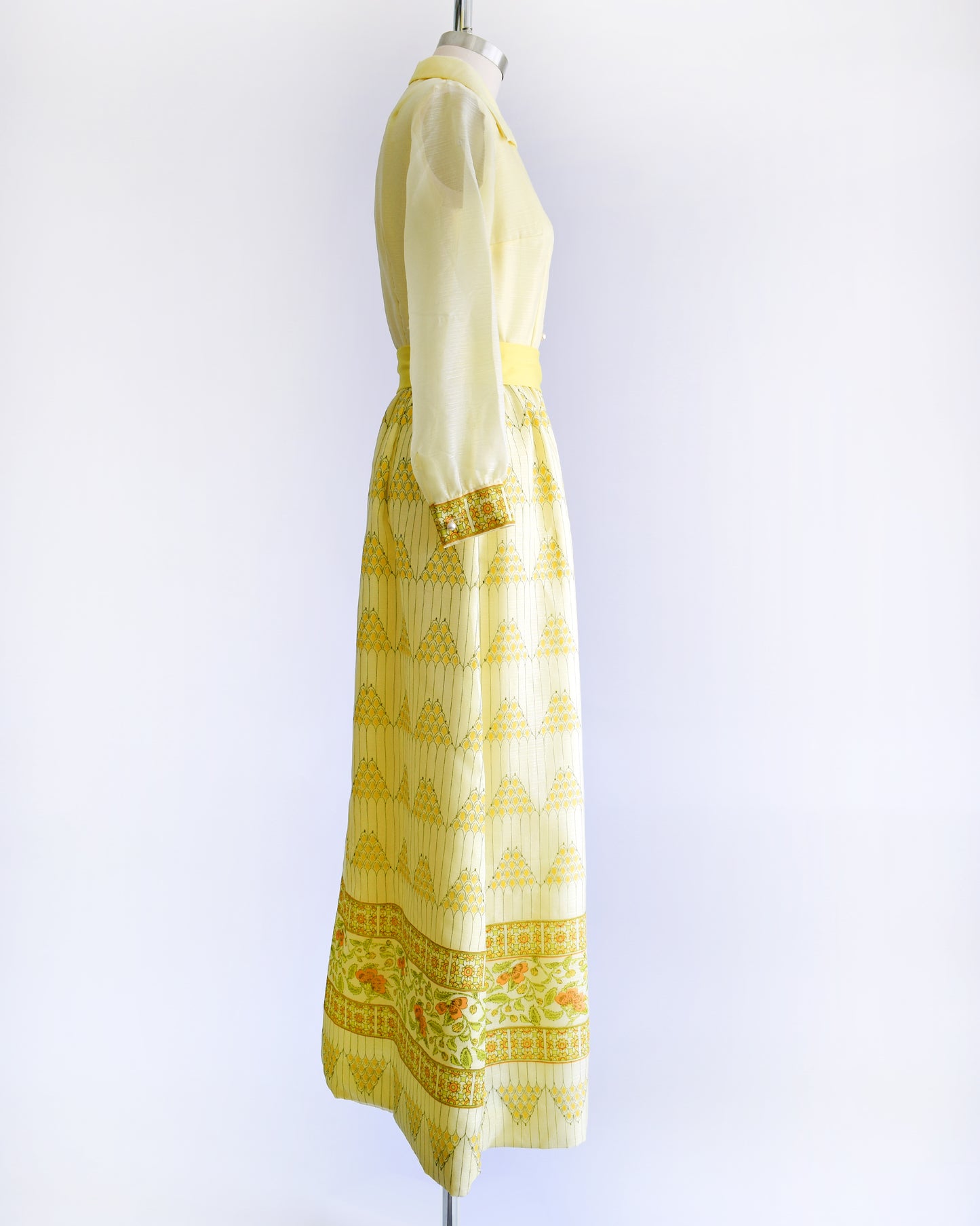 Side view of a yellow vintage maxi dress on a dress form. The dress features a collared neckline, semi sheer long sleeves, button down front, yellow sash belt, and a maxi skirt that has a pyramid and floral print.