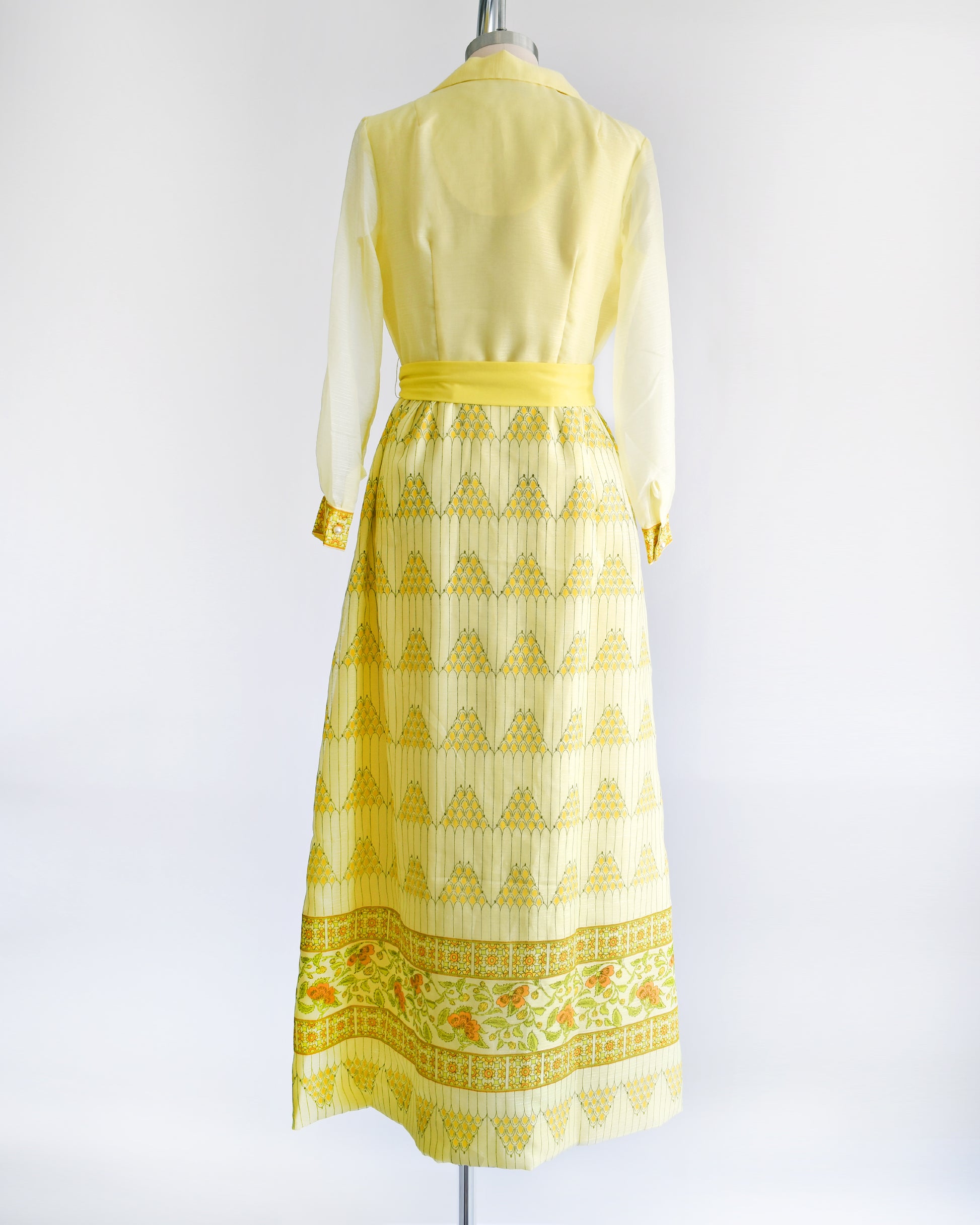 Back view of a yellow vintage maxi dress on a dress form. The dress features a collared neckline, semi sheer long sleeves, button down front, yellow sash belt, and a maxi skirt that has a pyramid and floral print.