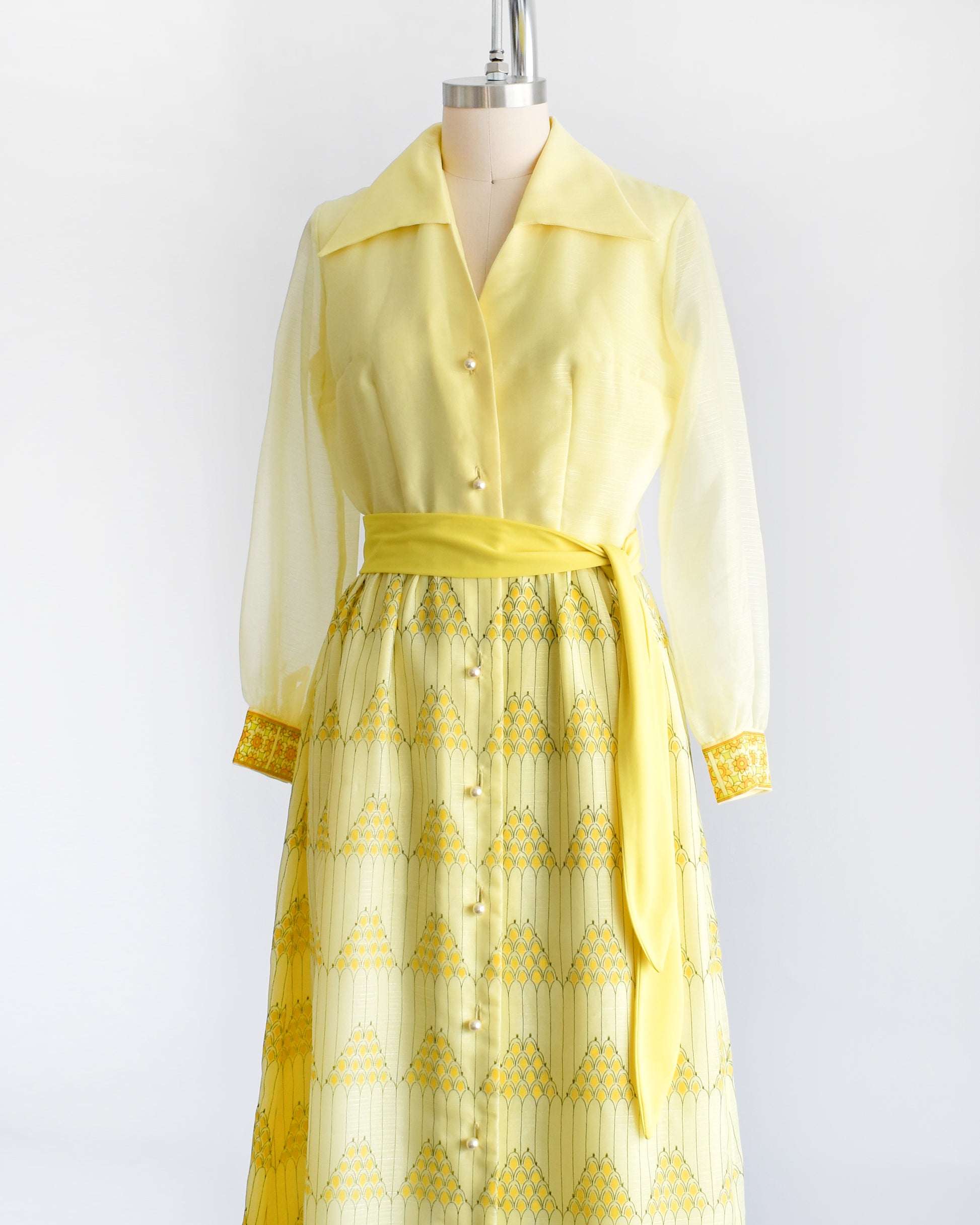 Side front view of a yellow vintage maxi dress on a dress form. The dress features a collared neckline, semi sheer long sleeves, button down front, yellow sash belt, and a maxi skirt that has a pyramid and floral print.
