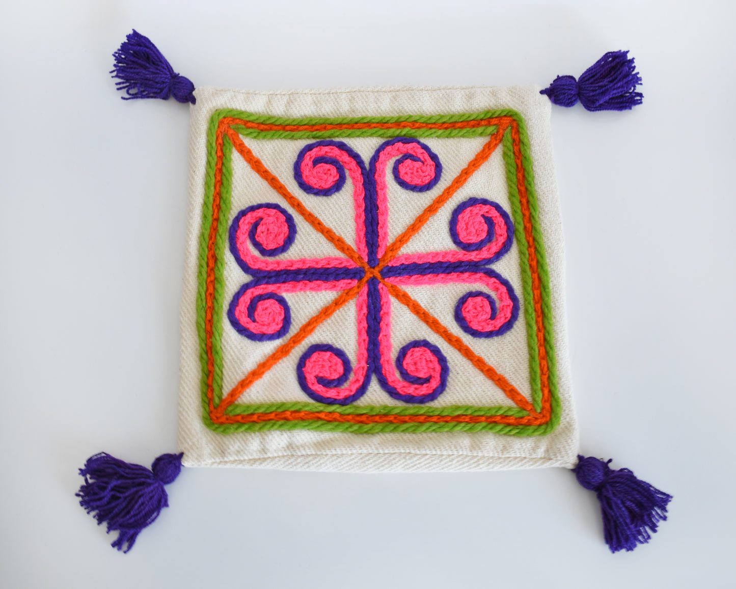 A vintage crewel embroidered pillow cover with purple tassels on a table.
