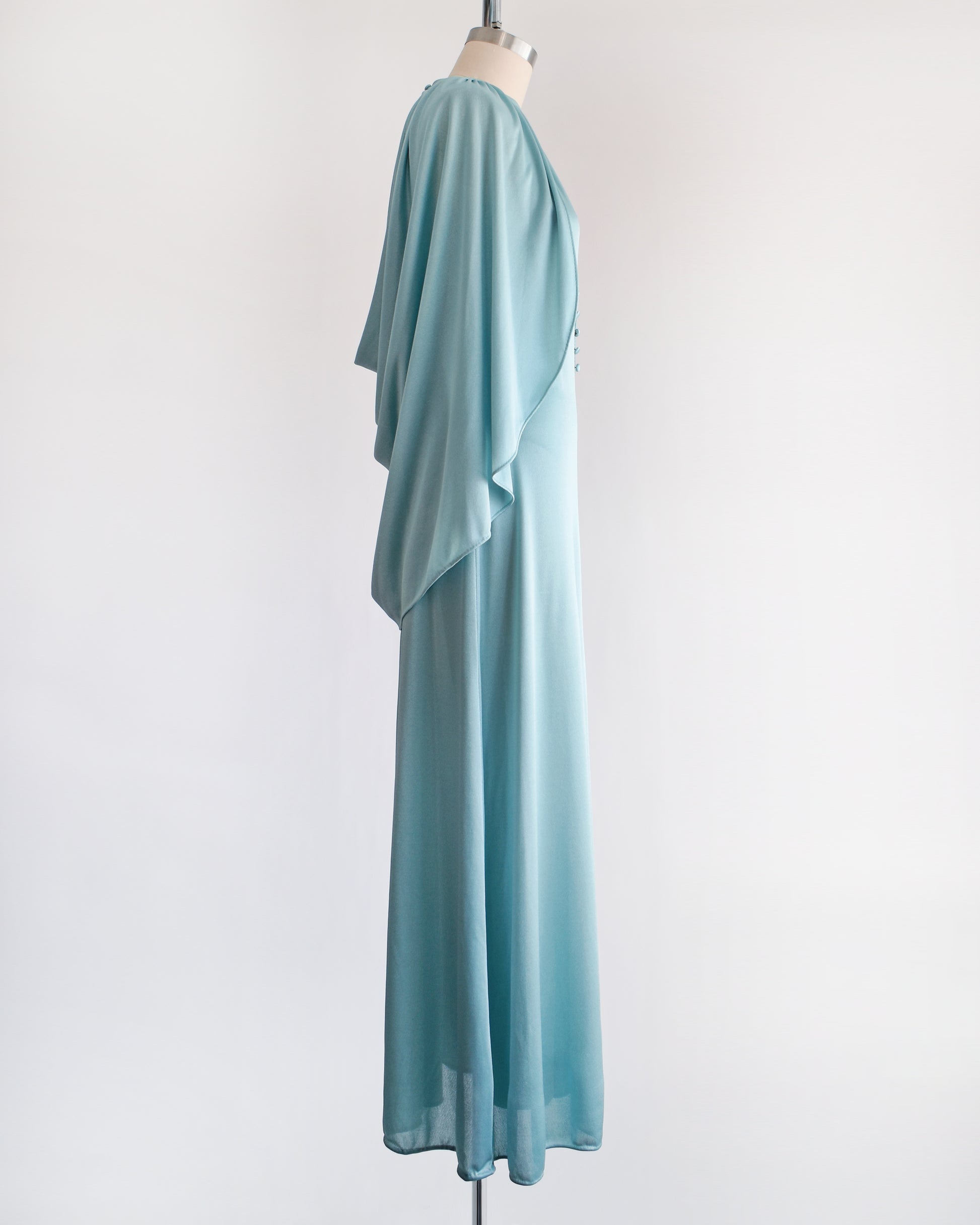 Side front view of a vintage 1970s seafoam blue caped maxi dress that has a triangle keyhole cutout and decorative buttons down the front