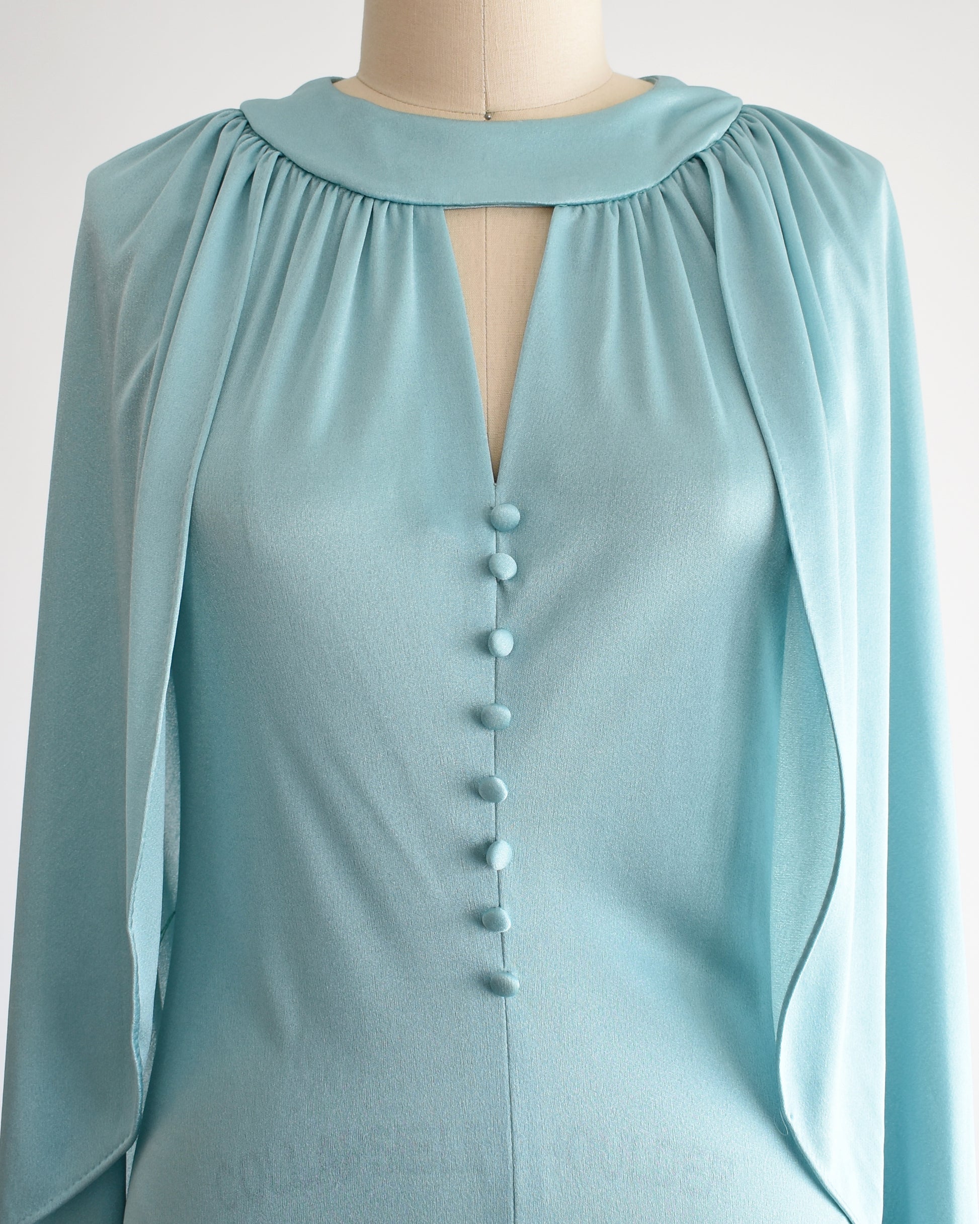 Close up of the bodice of a vintage 1970s seafoam blue caped maxi dress that has a triangle keyhole cutout and decorative buttons down the front