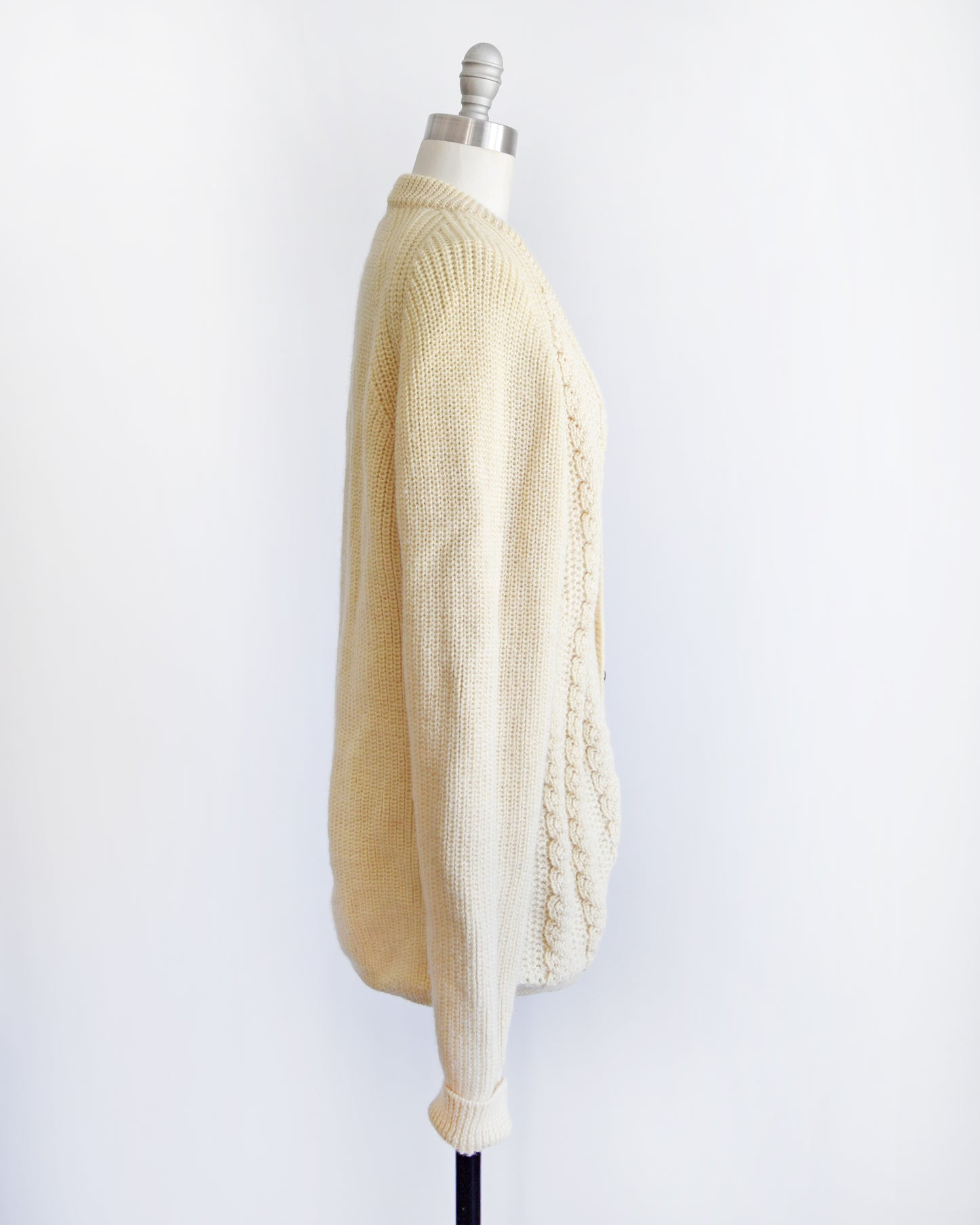 Side view of a vintage cream cable knit cardigan with dark buttons down the front. The cardigan is modeled on a dress form.