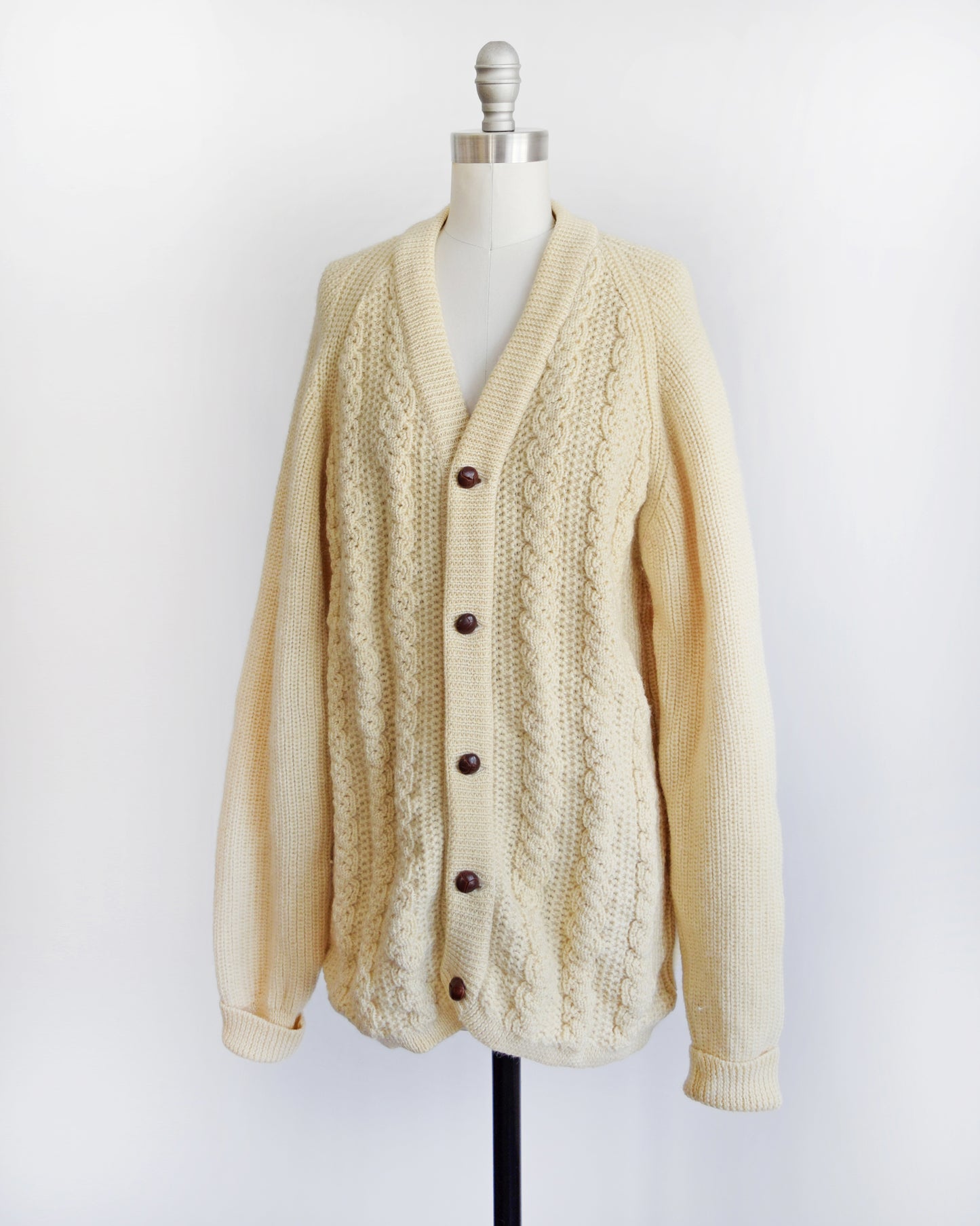 Side front view of a vintage cream cable knit cardigan with dark buttons down the front. The cardigan is modeled on a dress form.
