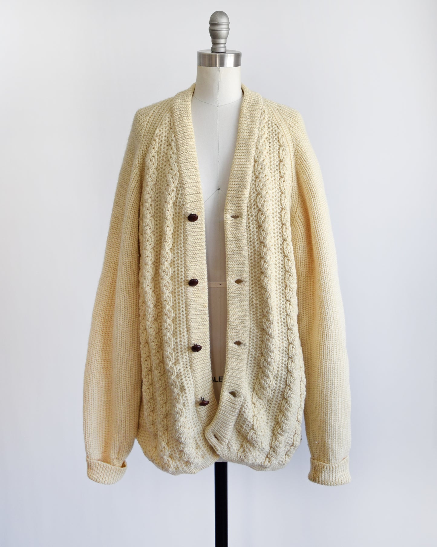 A vintage cream cable knit cardigan with dark buttons down the front. The cardigan is unbuttoned in this photo and is modeled on a dress form.