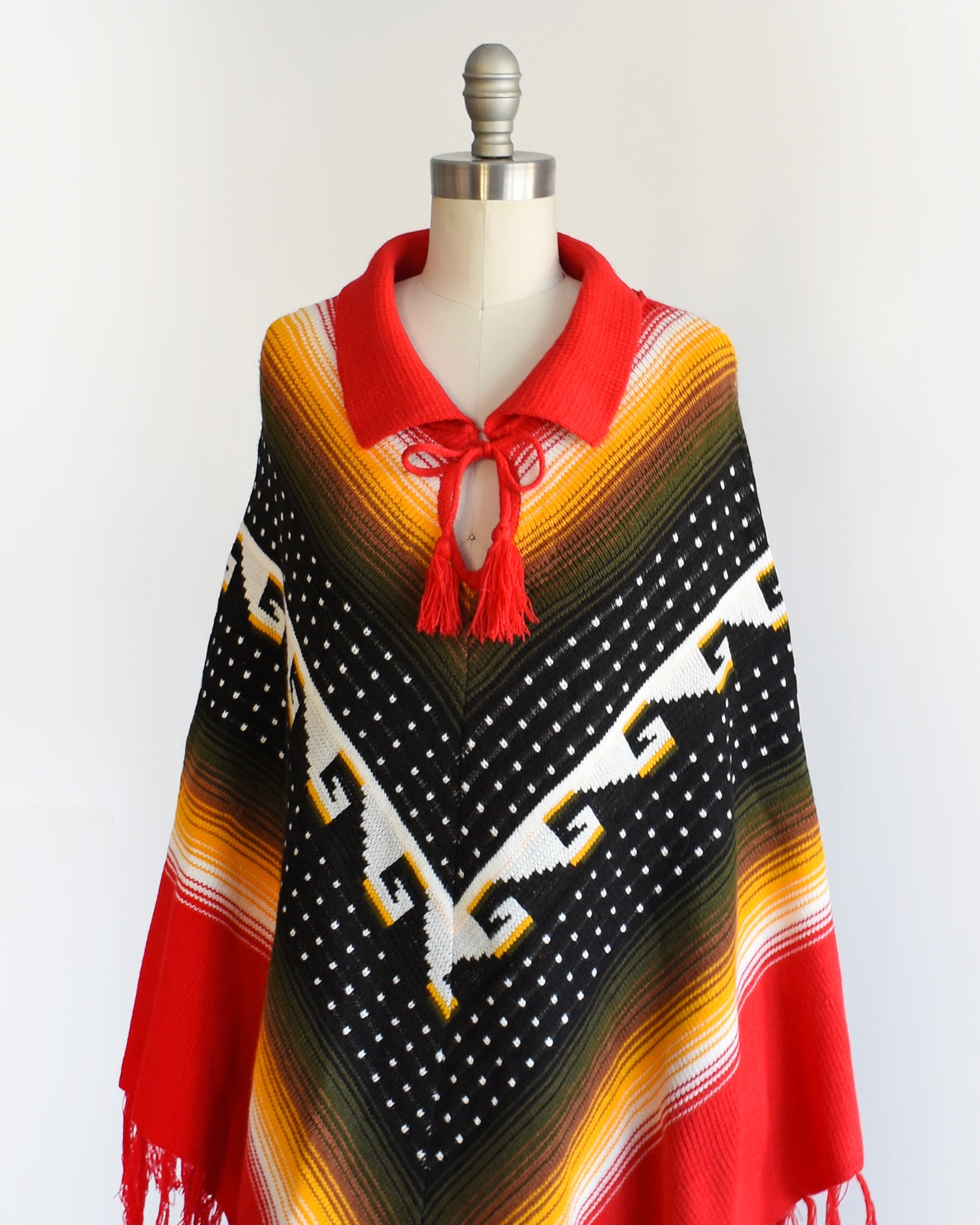 Side front view of a vintage 1970s chevron striped fringe poncho that has a folded over red collar with and two tassels that can be tied into a bow.