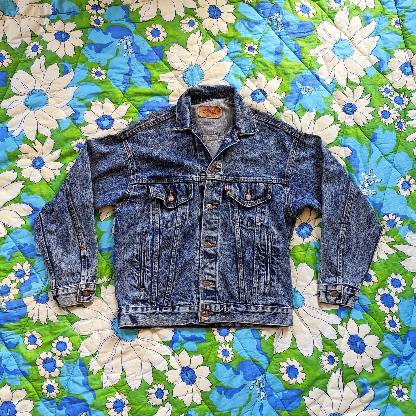 A flat lay photo of a vintage 80s Levi&#39;s blue jean denim jacket button up on a floral blanket