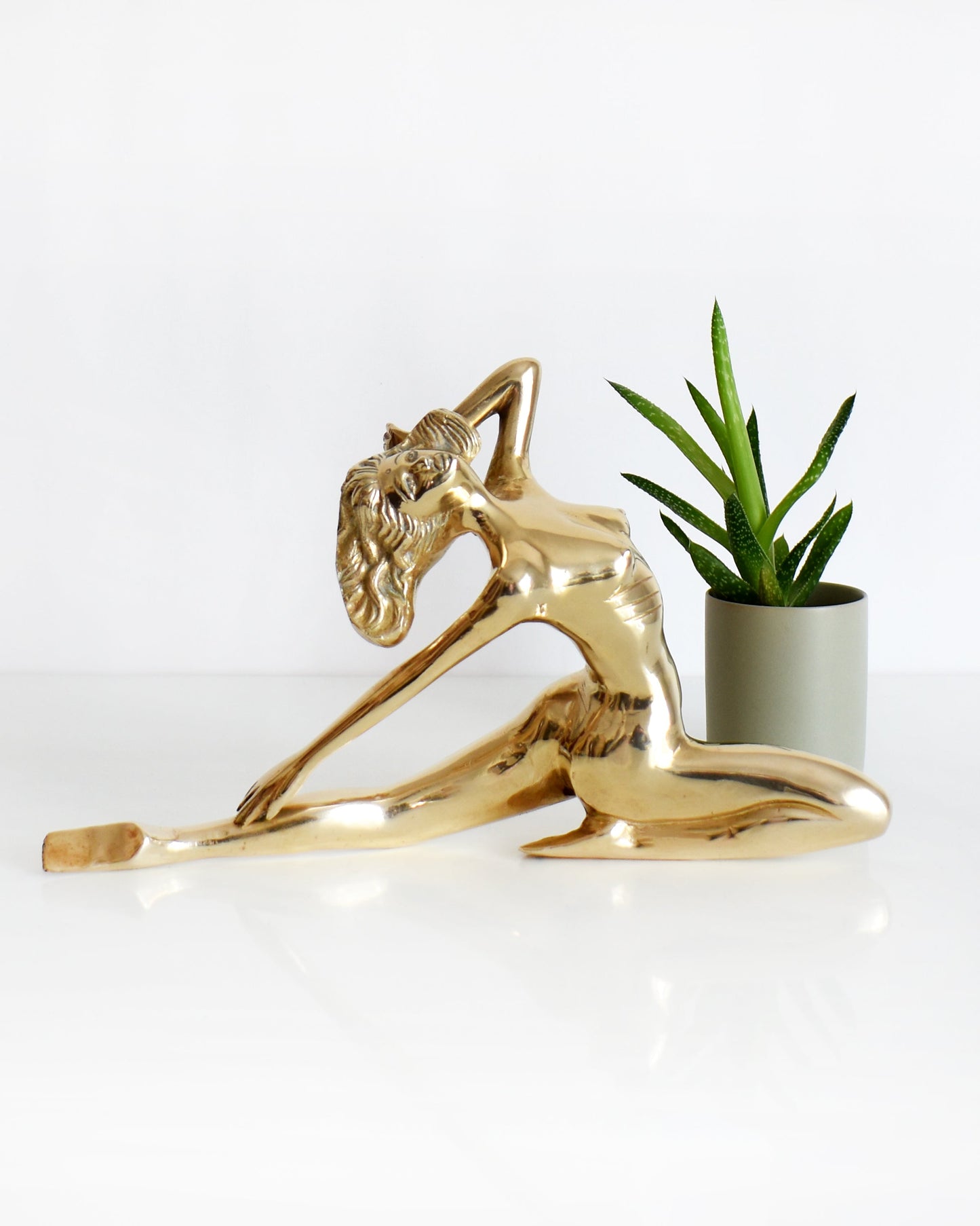 A vintage 1970s nude brass woman that is posed with one arm bent, another stretched behind her, and one leg also stretched out and the other leg is bent at the knee. There is a small potted plant in the background