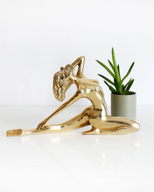 A vintage 1970s nude brass woman that is posed with one arm bent, another stretched behind her, and one leg also stretched out and the other leg is bent at the knee.  There is a small potted plant in the background