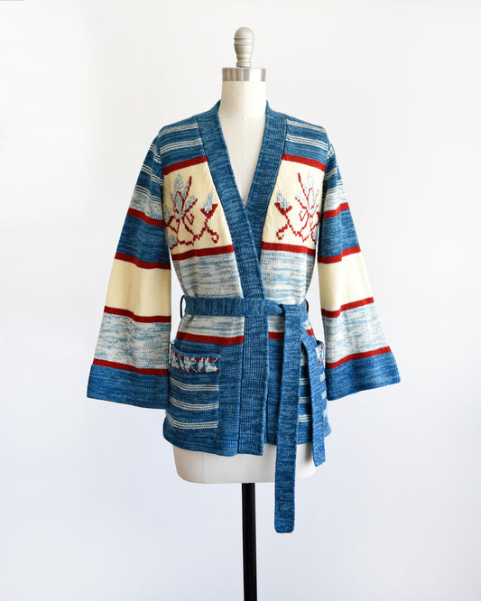 a vintage 1970s blue floral space dye belted cardigan that has dark and light blue space dye knit with red and cream white stripes and large blue and red flowers on the front of the sweater.