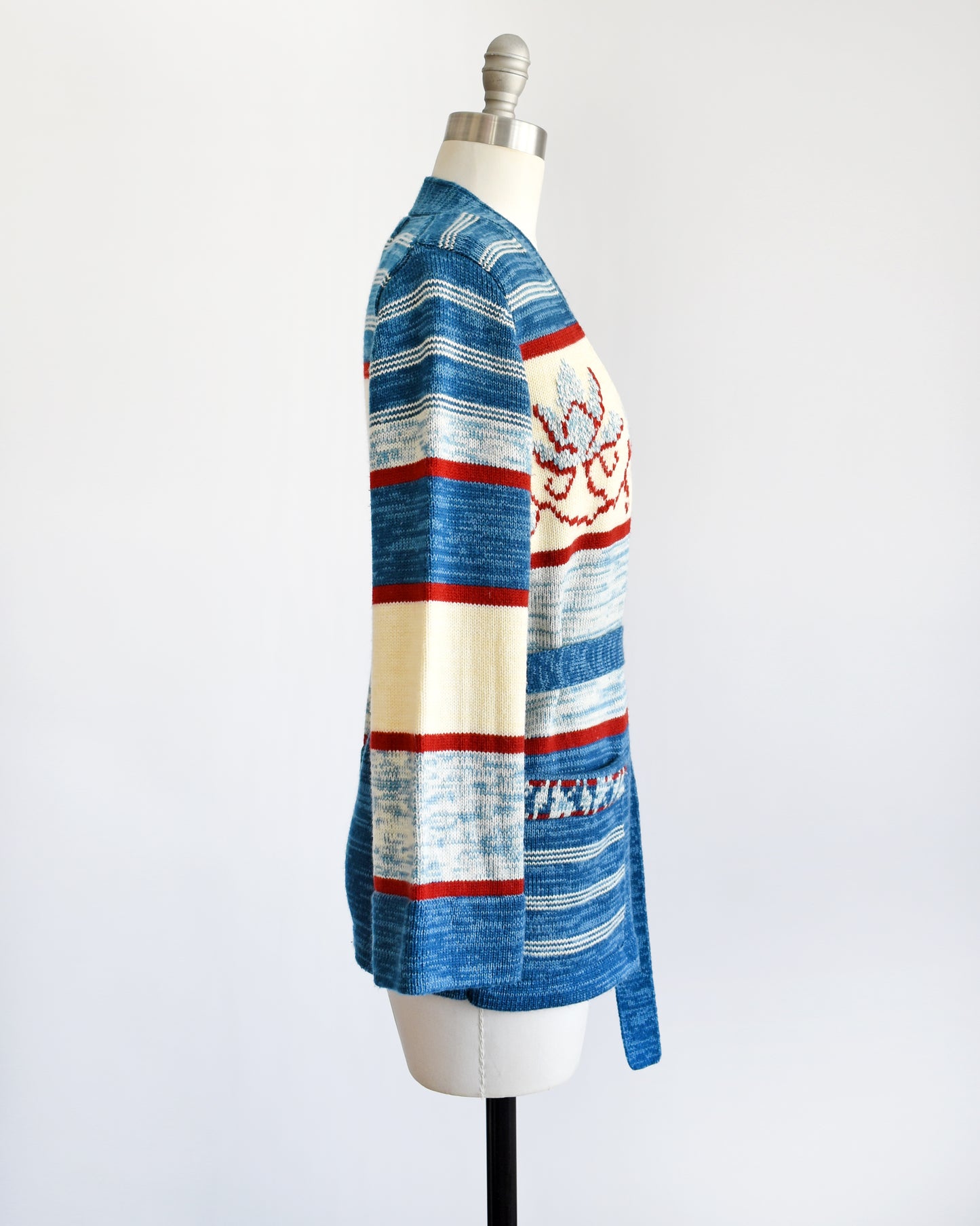 side view of a vintage 1970s blue floral space dye belted cardigan that has dark and light blue space dye knit with red and cream white stripes and large blue and red flowers on the front of the sweater.