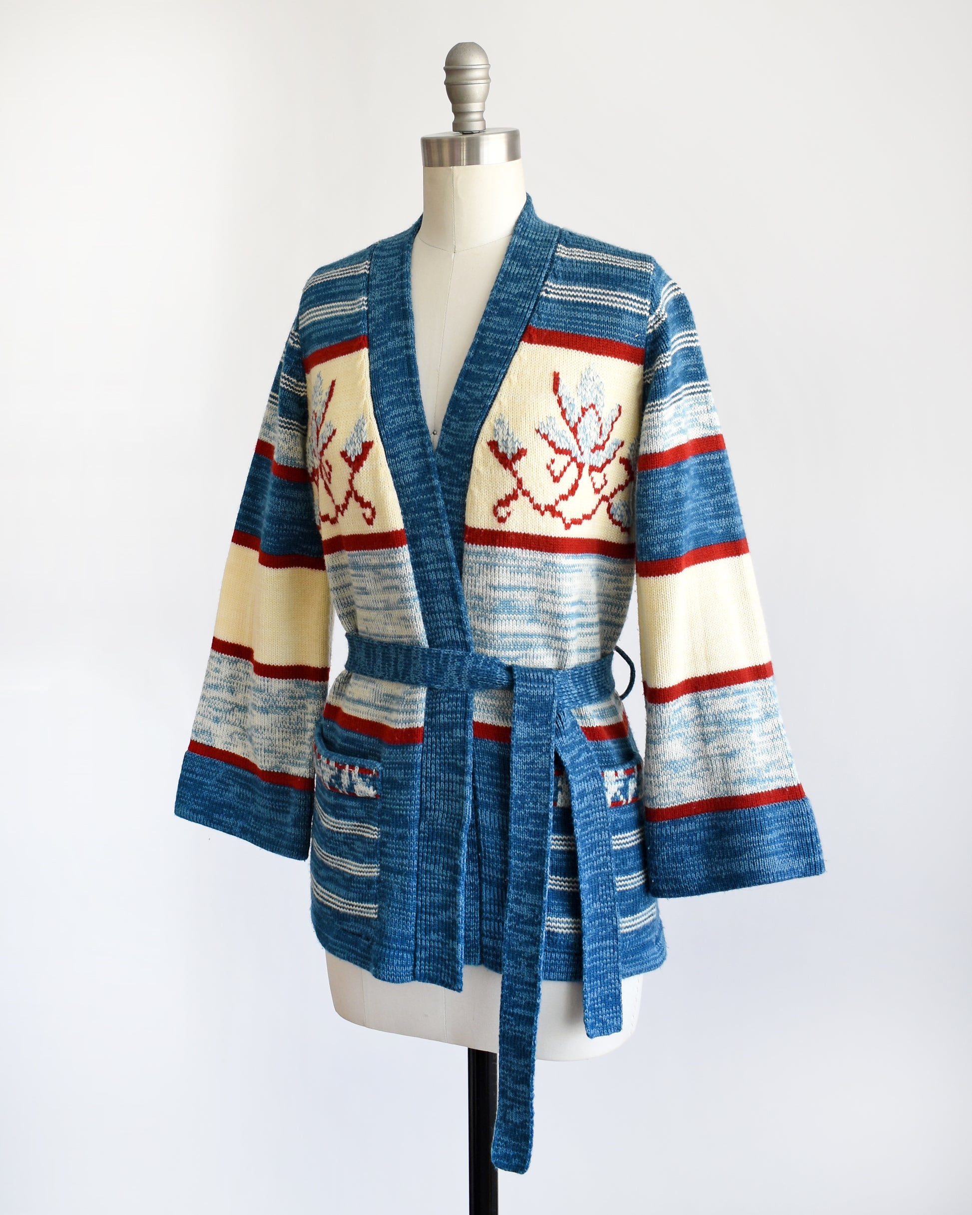 side front view of a vintage 1970s blue floral space dye belted cardigan that has dark and light blue space dye knit with red and cream white stripes and large blue and red flowers on the front of the sweater.
