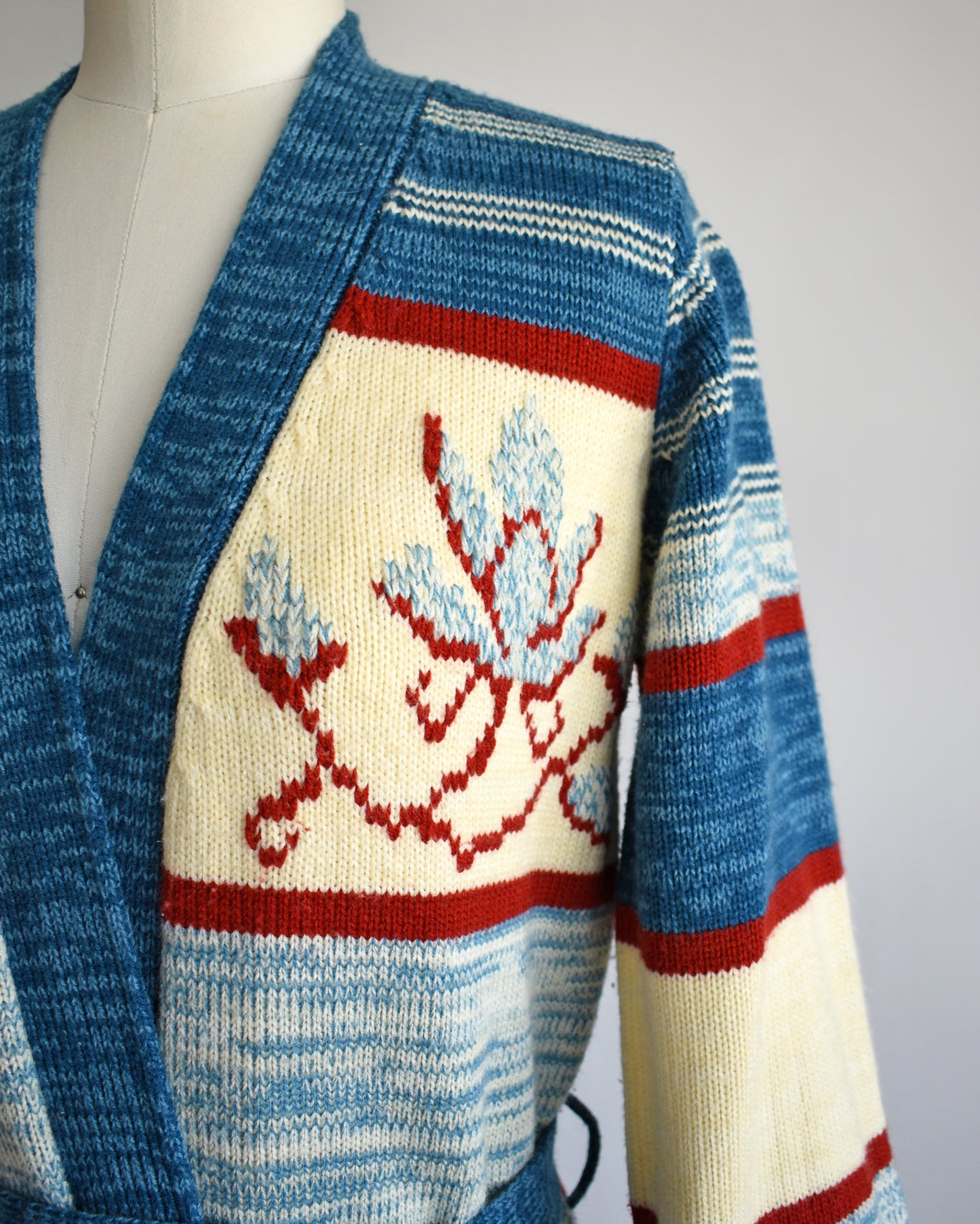 close up of a vintage 1970s blue floral space dye belted cardigan that has dark and light blue space dye knit with red and cream white stripes and large blue and red flowers on the front of the sweater.