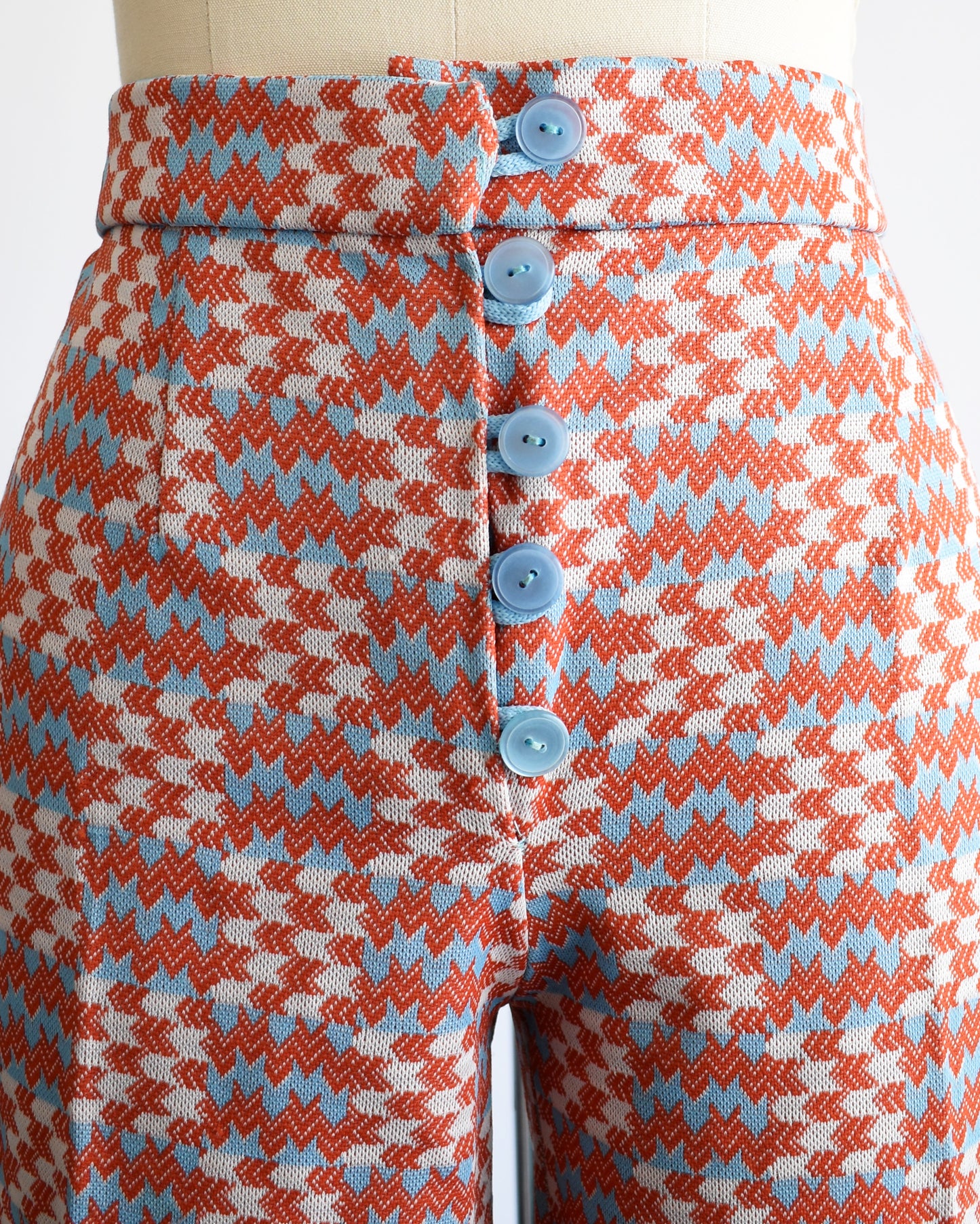 close up of the a pair of vintage 1970s wide leg pants feature a vibrant print with dark orange, white, and light blue arrows pointing in various directions down the legs. The pants have 4 light blue buttons on the front