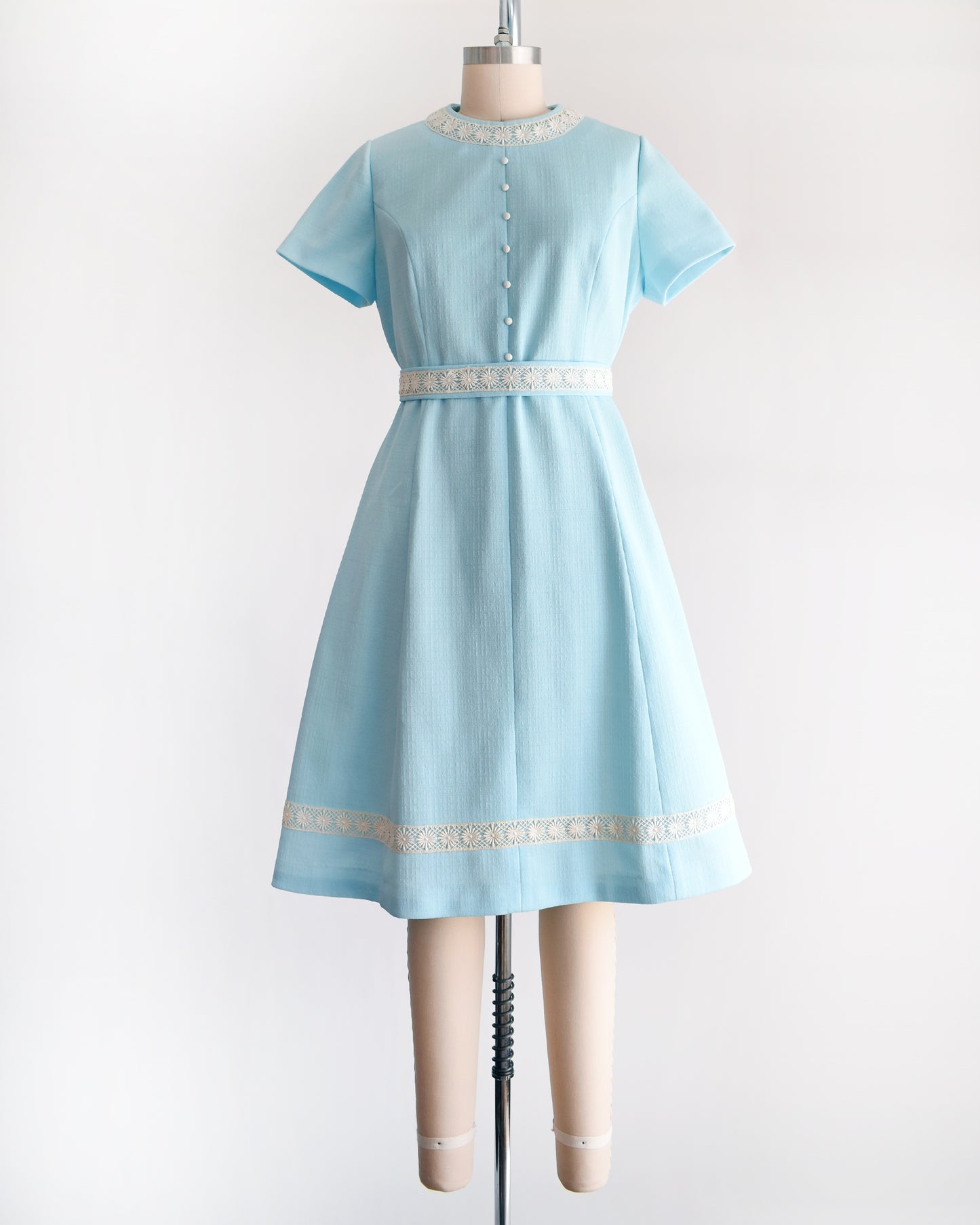 a late 1960s early 1970s vintage mod dress that is light blue and has cream floral lace trim around the collar, belt, and hem