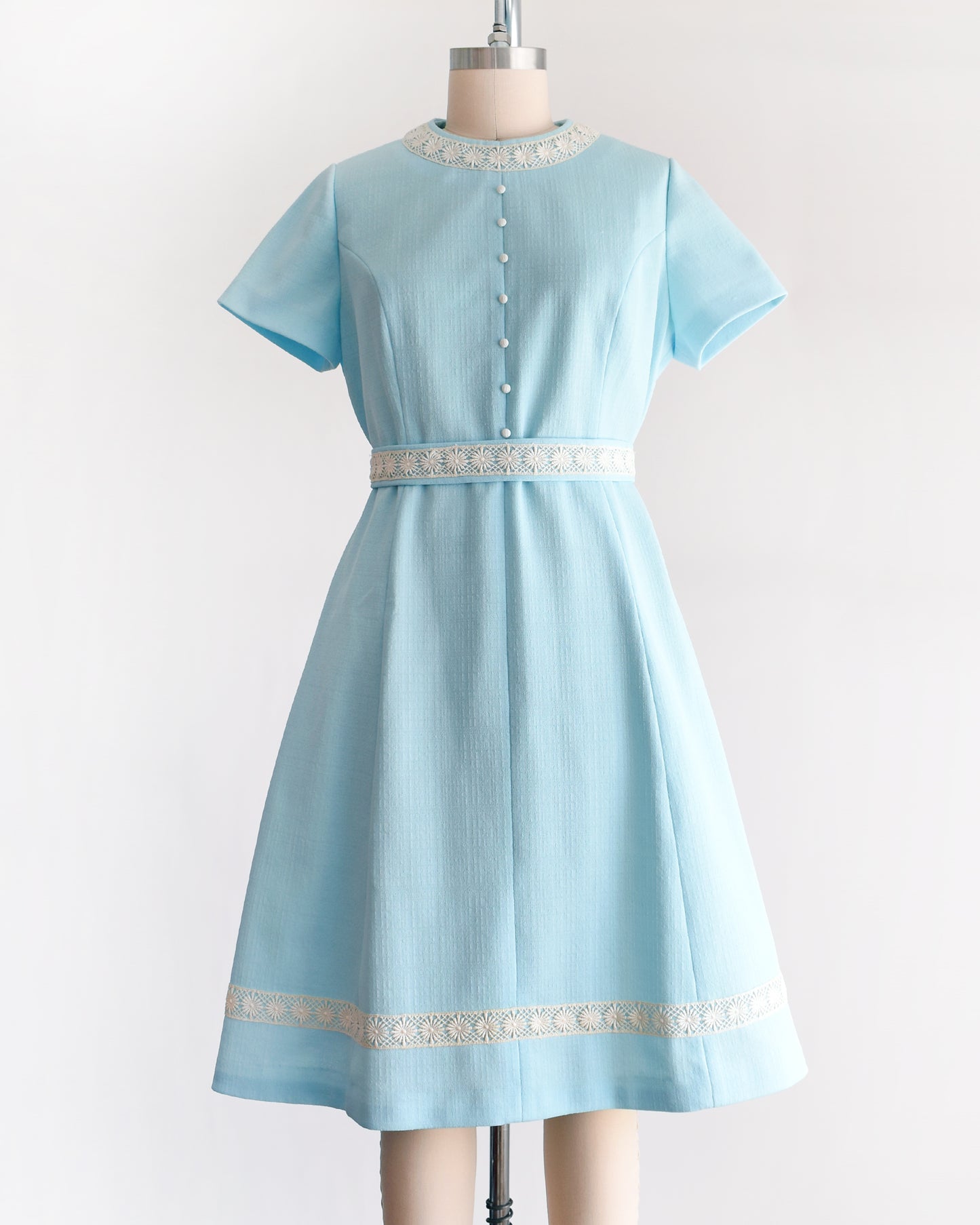 a late 1960s early 1970s vintage mod dress that is light blue and has cream floral lace trim around the collar, belt, and hem