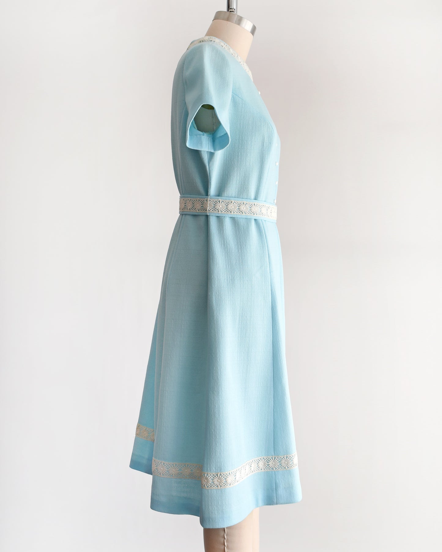 side view of a late 1960s early 1970s vintage mod dress that is light blue and has cream floral lace trim around the collar, belt, and hem