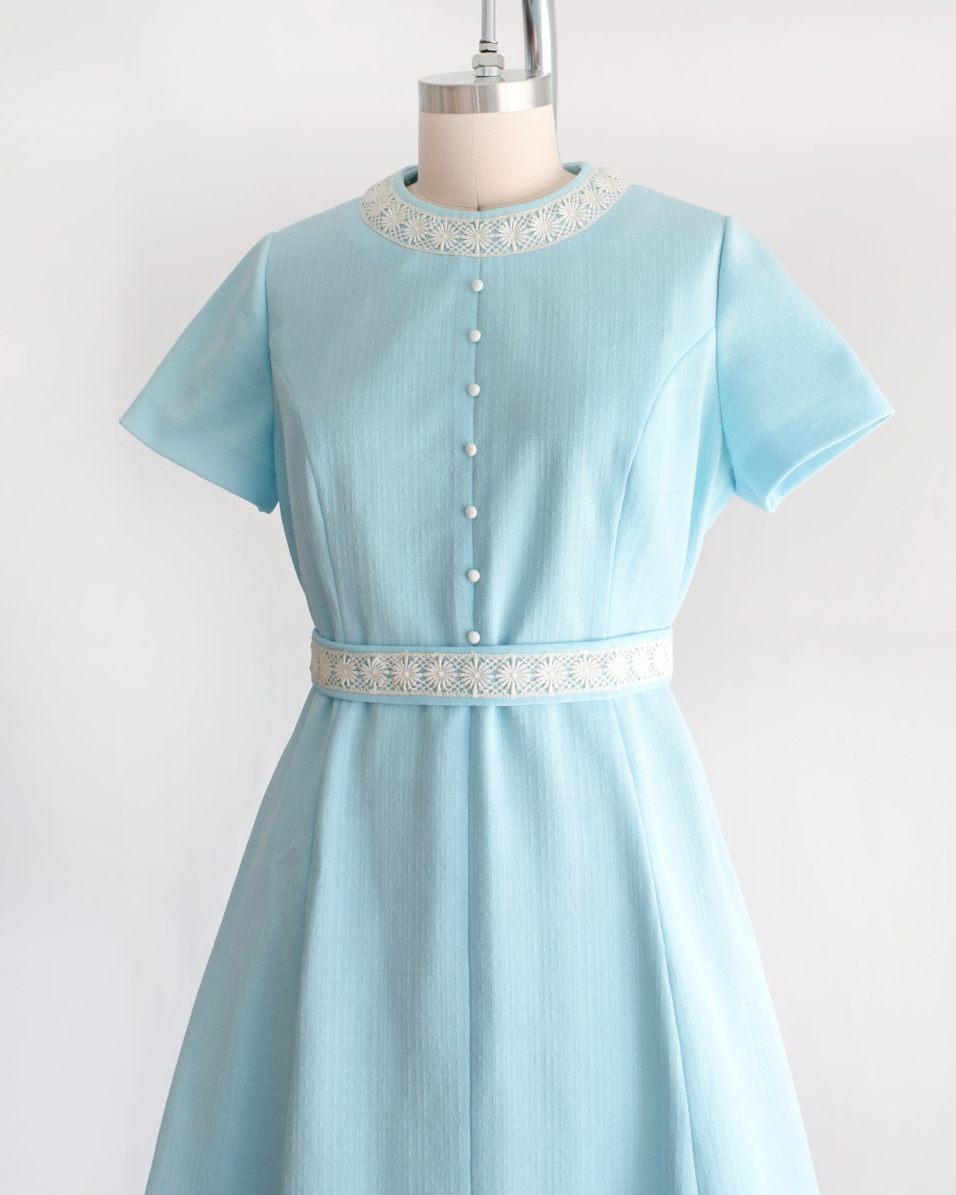 side front view of a late 1960s early 1970s vintage mod dress that is light blue and has cream floral lace trim around the collar, belt, and hem