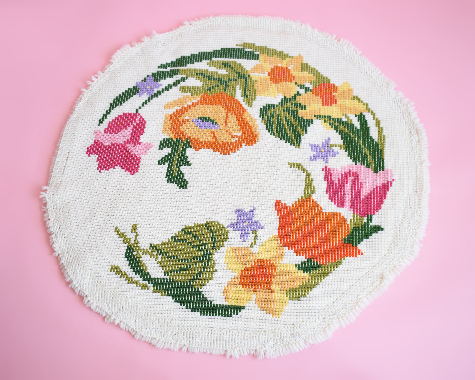 Back view of a vintage round floral latch hook rug that has cream white shaggy acrylic with a colorful floral motif in pinks, orange, yellow, purple and greens. The rug is on a pink background.