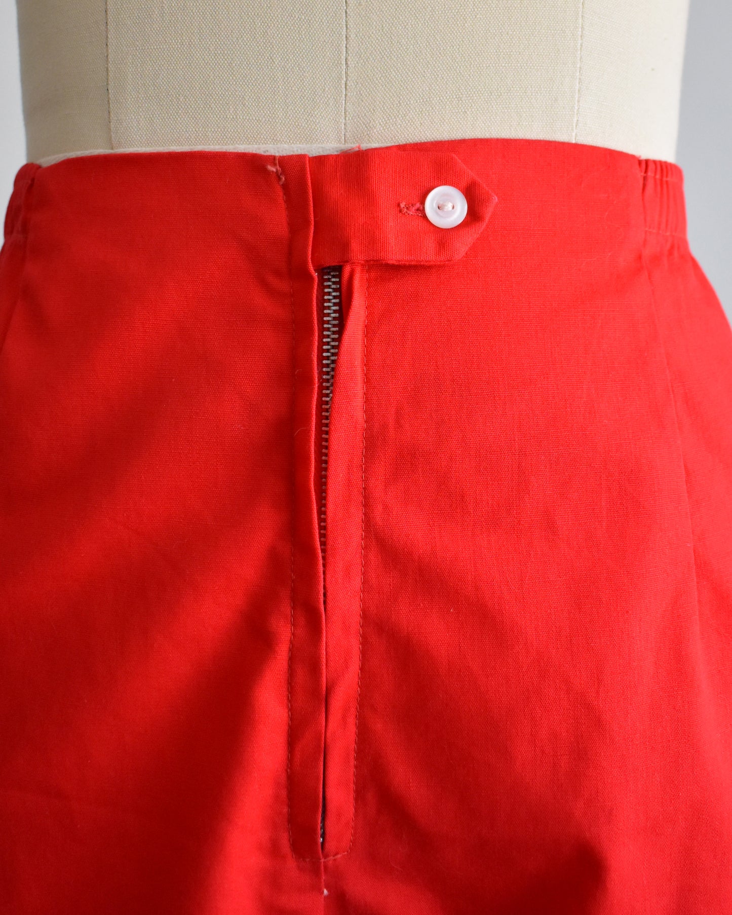 close up of the waist, tab button, and metal zipper on a pair of vintage 1960s red shorts on a mannequin against a white background