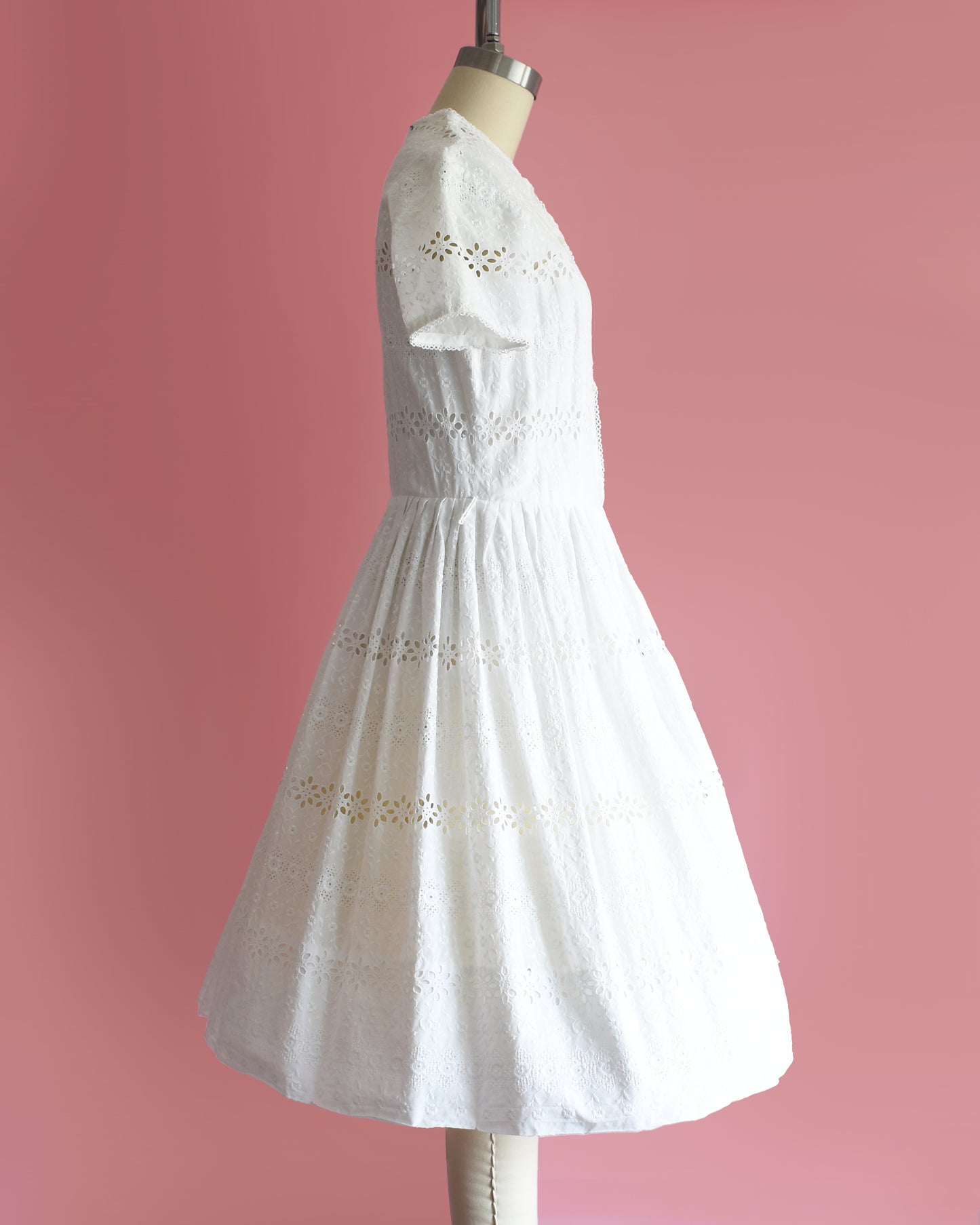side view of a vintage 1960s L’Aiglon eyelet white dress on a mannequin against a pink background
