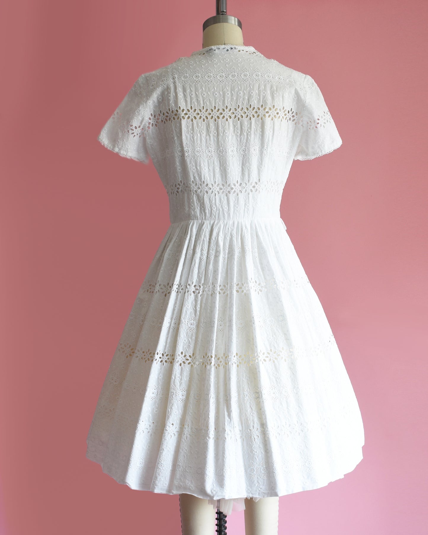 back view of a vintage 1960s L’Aiglon eyelet white dress on a mannequin against a pink background
