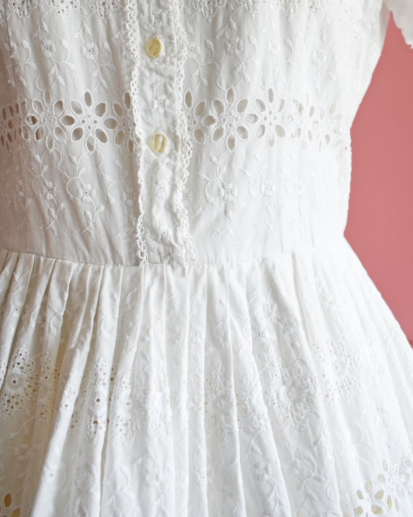 close up of the waist of a vintage 1960s L’Aiglon eyelet white dress on a mannequin against a pink background