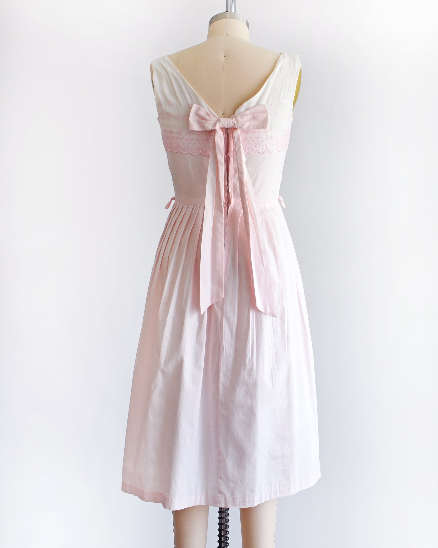 back view of a vintage 1950s pink and white embroidered dress that has a back bow on a mannequin against a white background