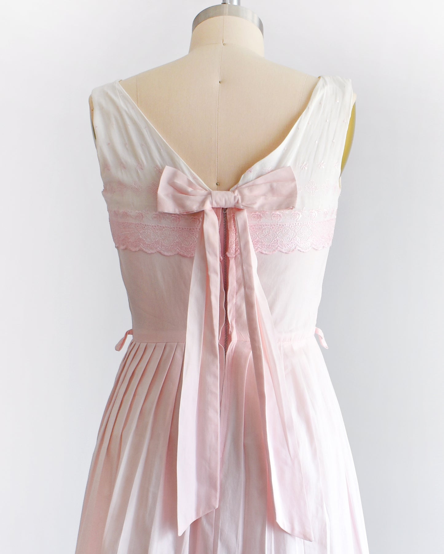 close up of the pink bow on the back of a vintage 1950s pink and white embroidered dress on a mannequin against a white background