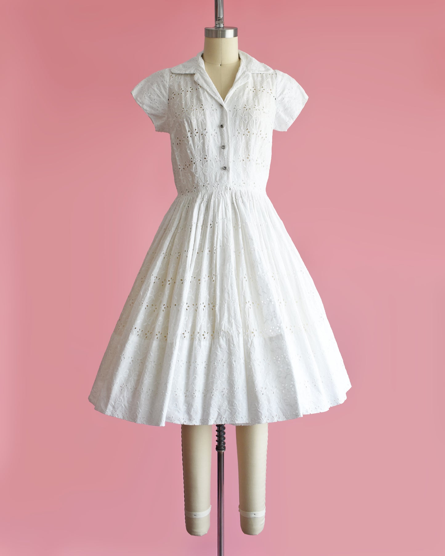 a vintage 1950s white eyelet dress with rhinestone buttons on a mannequin against a pink background