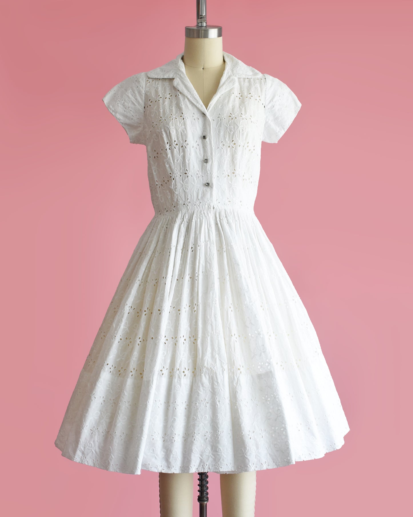 a vintage 1950s white eyelet dress with rhinestone buttons on a mannequin against a pink background