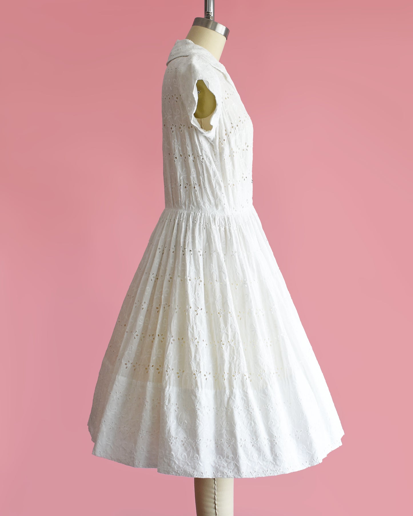 side view of a vintage 1950s white eyelet dress with rhinestone buttons on a mannequin against a pink background
