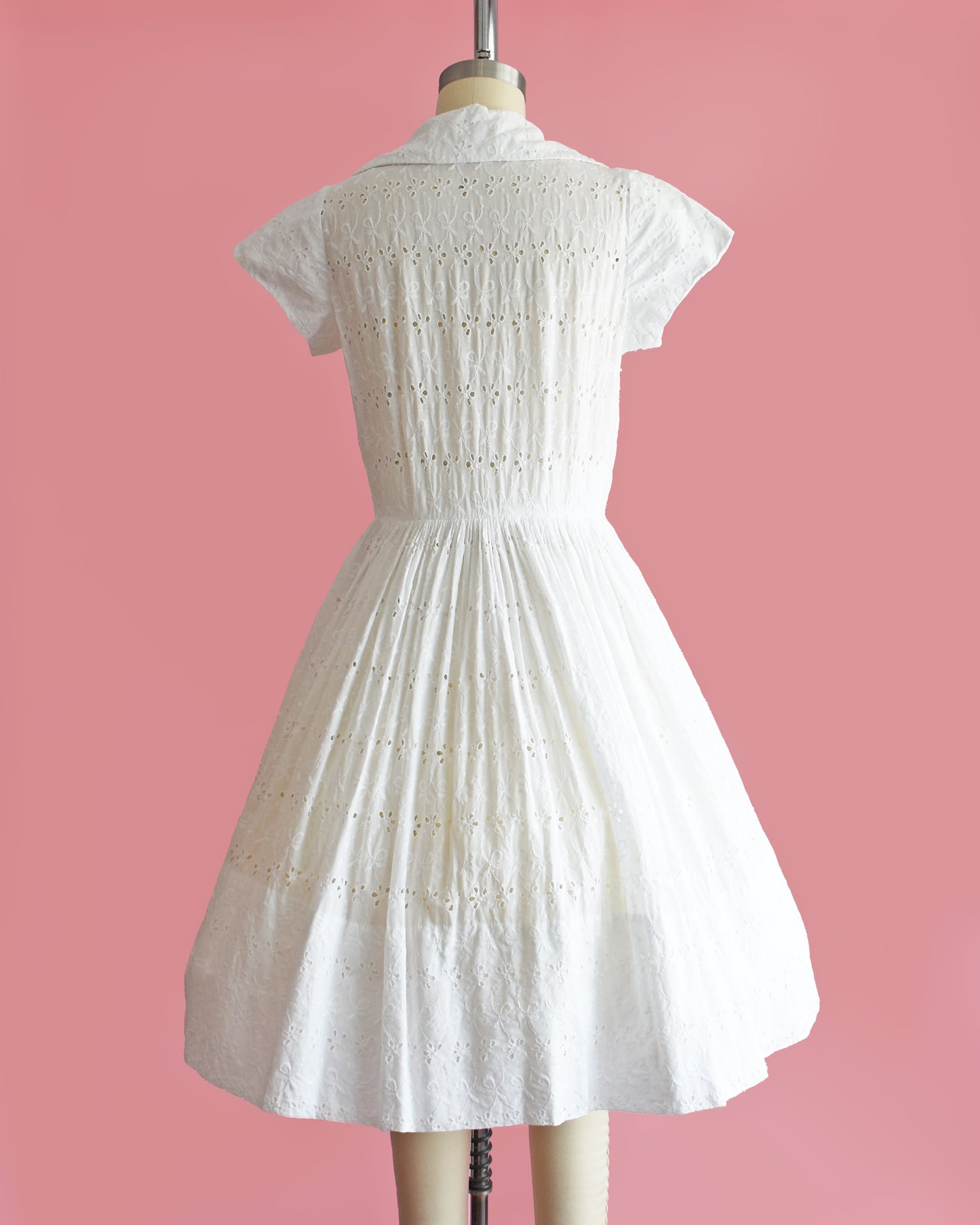 back view of a vintage 1950s white eyelet dress with rhinestone buttons on a mannequin against a pink background