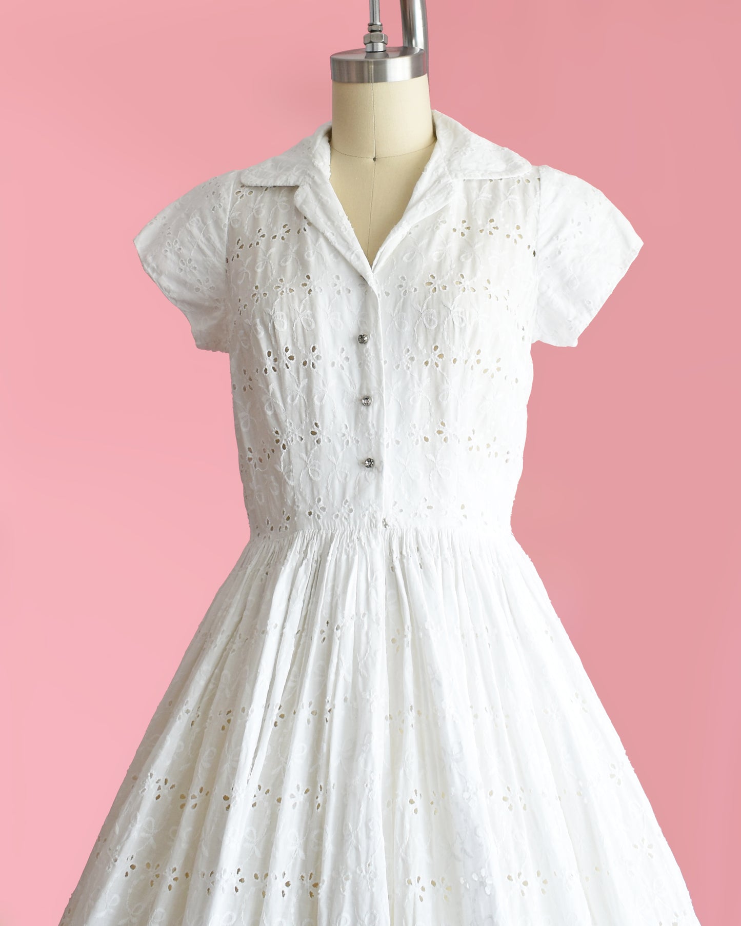 side front view of a vintage 1950s white eyelet dress with rhinestone buttons on a mannequin against a pink background