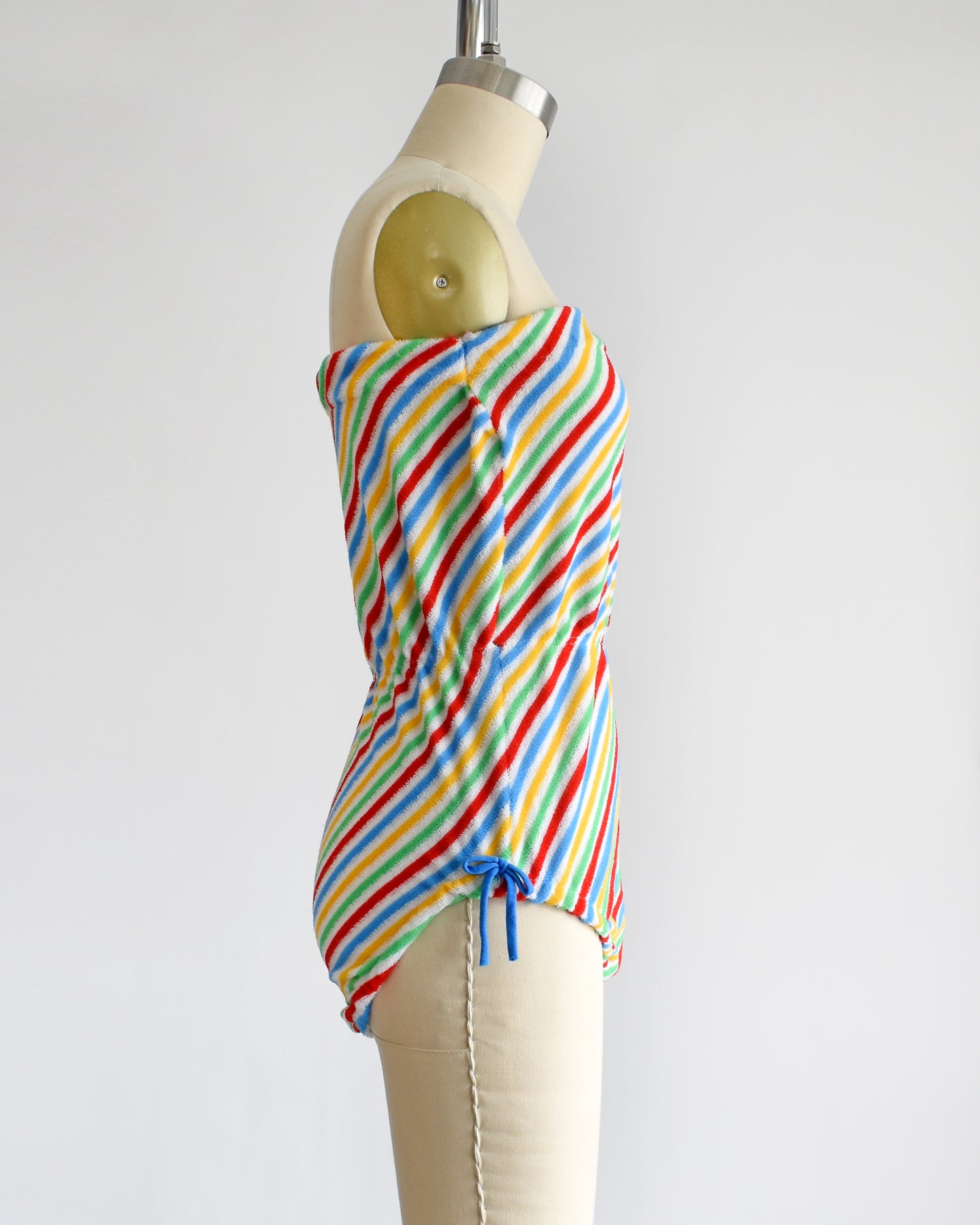 side view of a vintage 1970s romper that is white with red, green, yellow, and blue diagonal stripes and blue bows on the hips