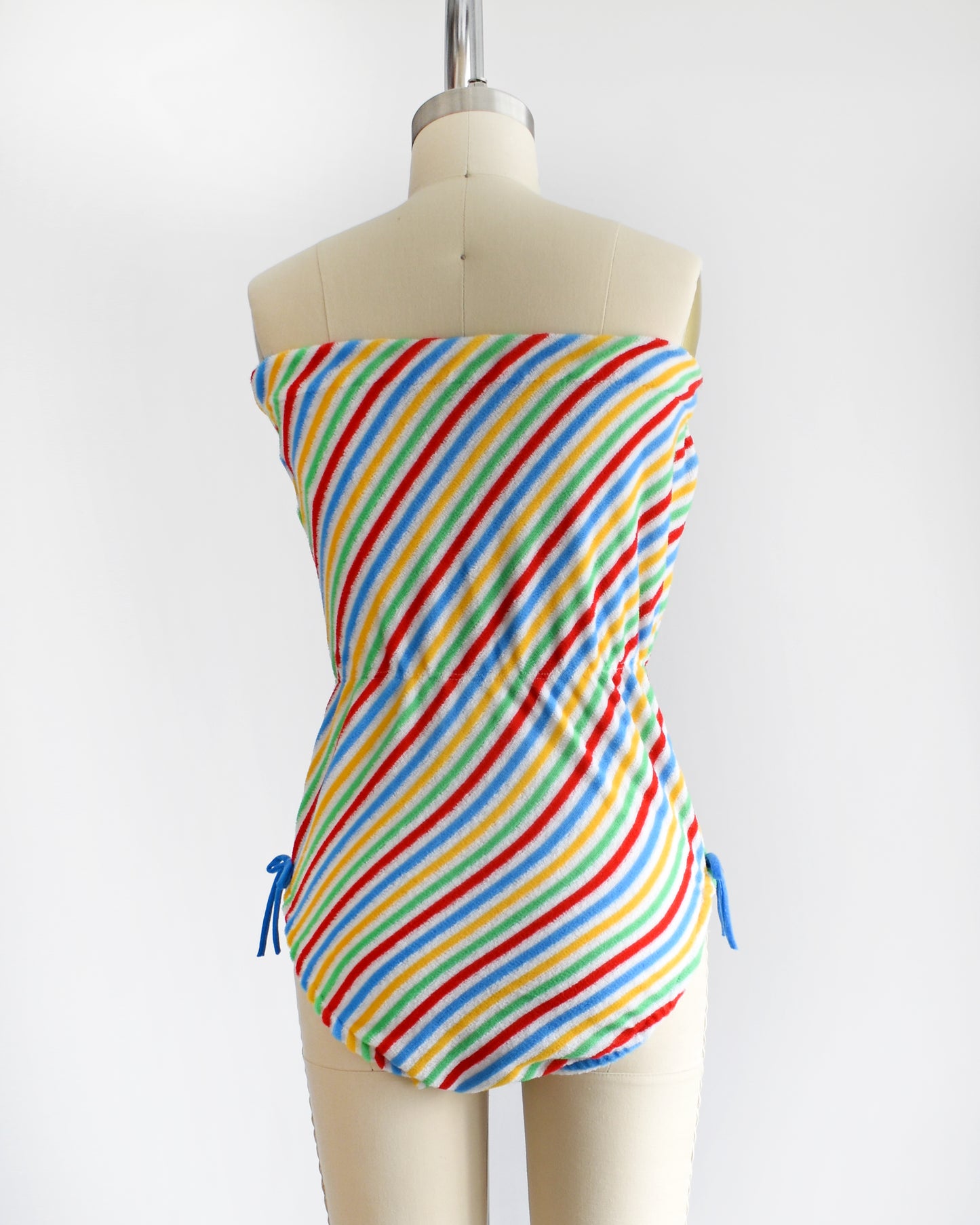 back view of a vintage 1970s romper that is white with red, green, yellow, and blue diagonal stripes and blue bows on the hips