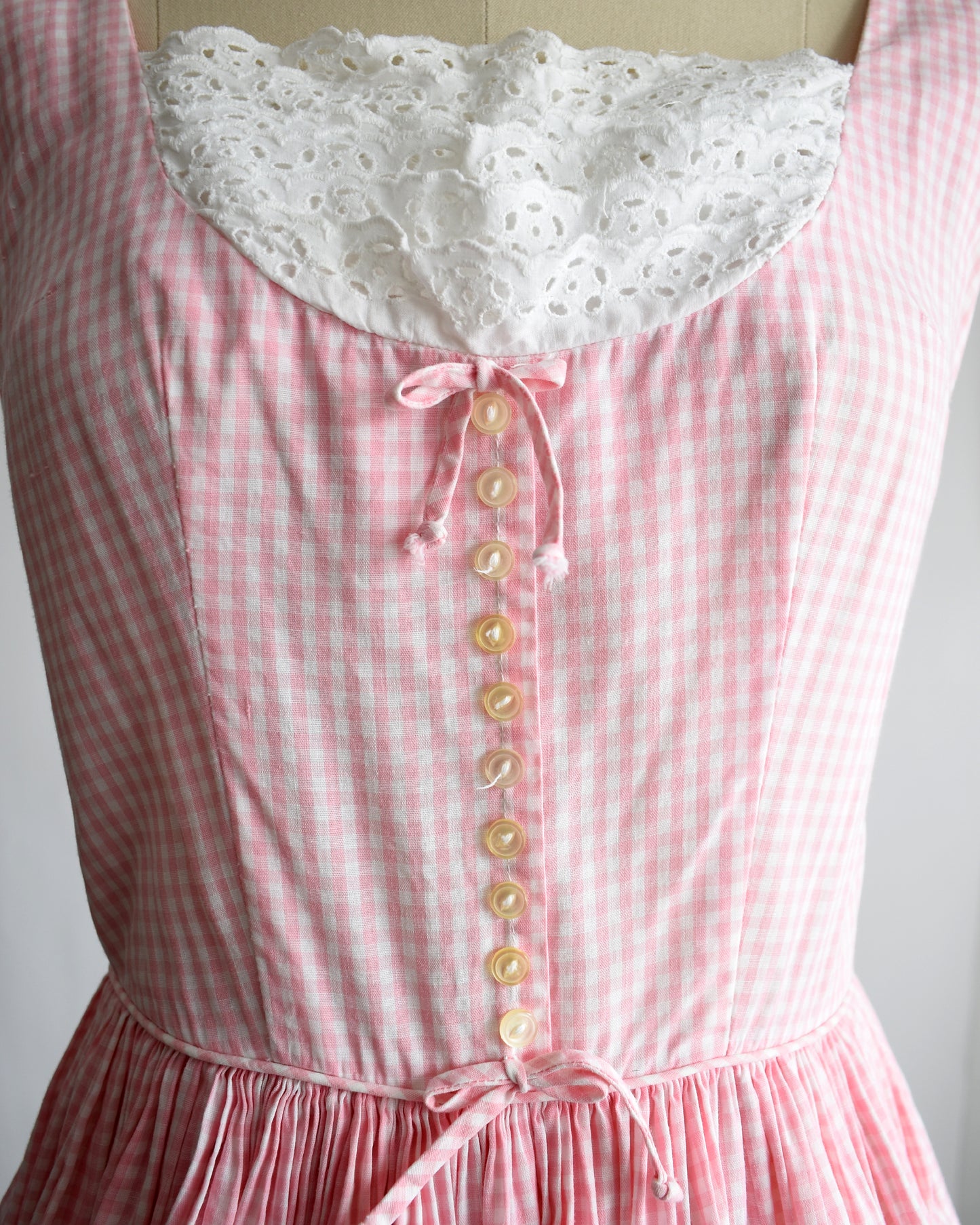 close up of a vintage 1950s pink and white gingham dress with a white lace bib on a mannequin against a white background