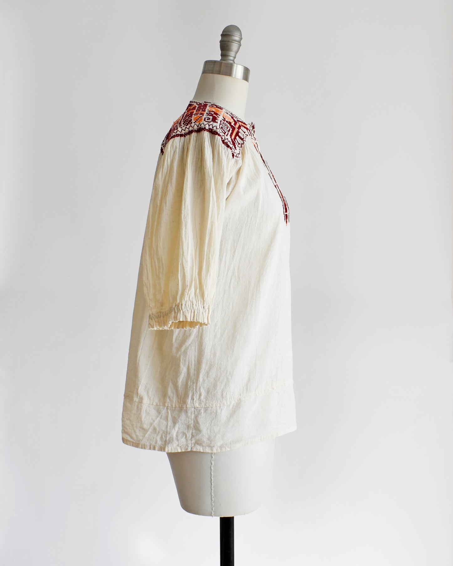 Side view of a vintage 60s cream crinkled cotton top with a burgundy and orange embroidered floral and what looks like llamas motif across the shoulders and down the front. The top is on a dress form.
