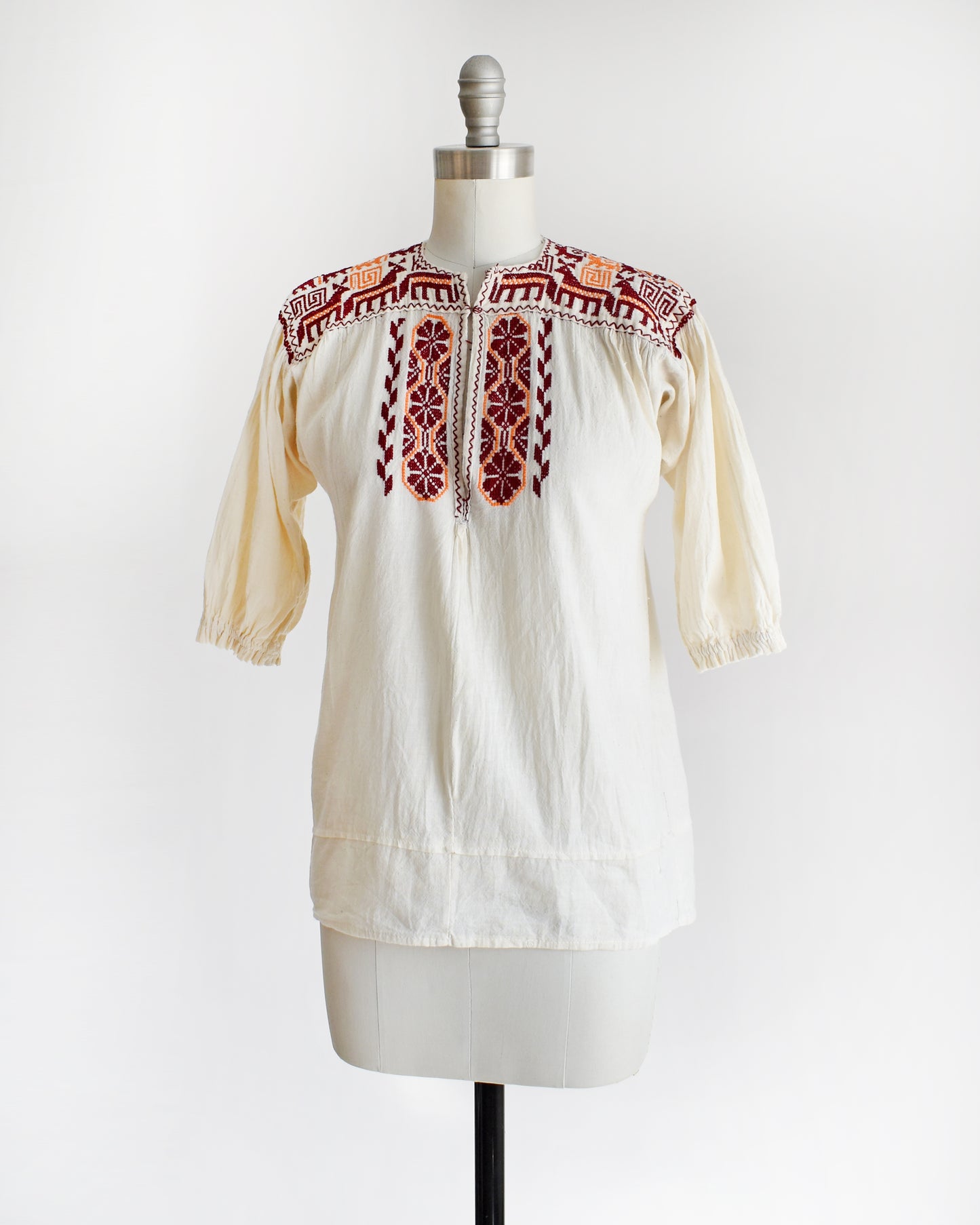 Side front view of a vintage 60s cream crinkled cotton top with a burgundy and orange embroidered floral and what looks like llamas motif across the shoulders and down the front. The top is on a dress form.