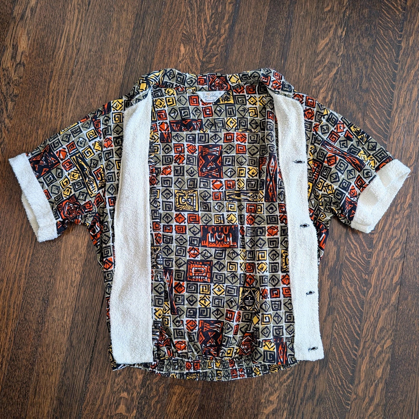 a vintage 1960s short-sleeve cabana shirt with geometric pattern which is unbuttoned and open on a wooden floor.