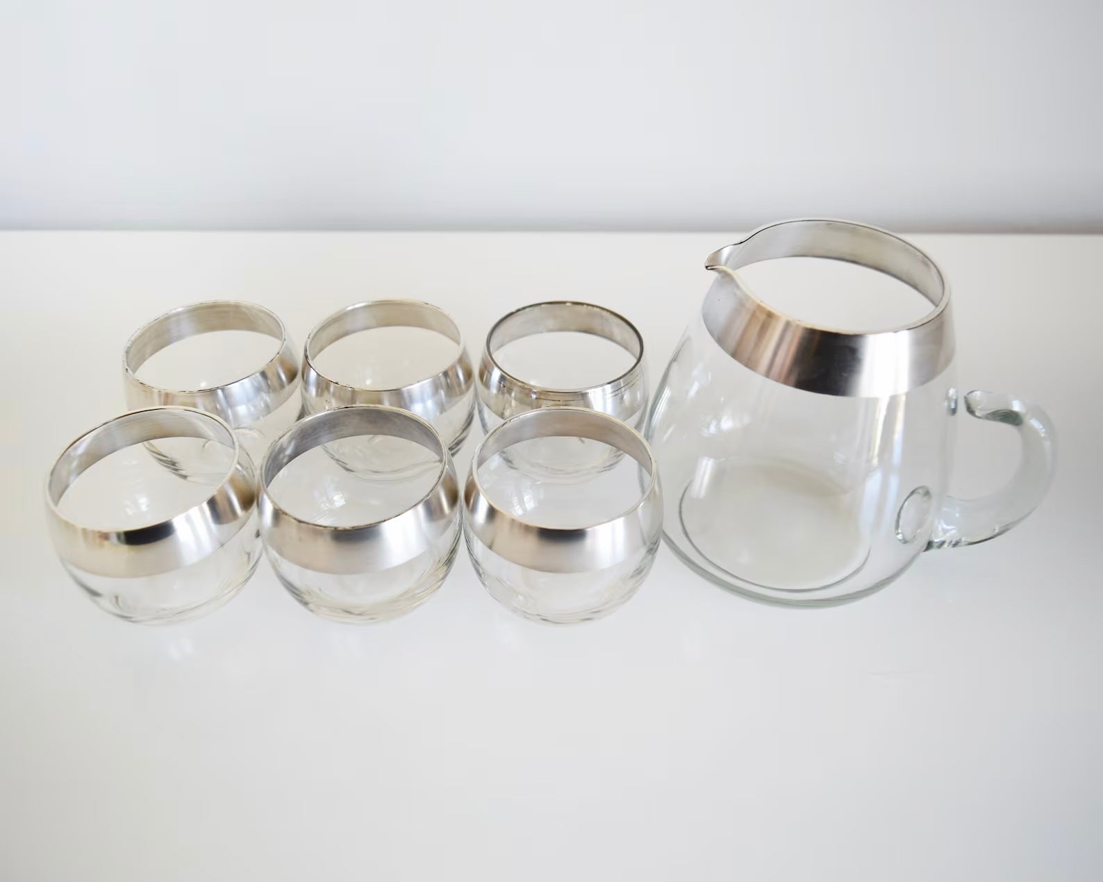 An overhead shot of a vintage Dorothy Thorpe cocktail set that has a set of six silver rimmed rounded roly poly glasses with a matching silver rim pitcher with handle.