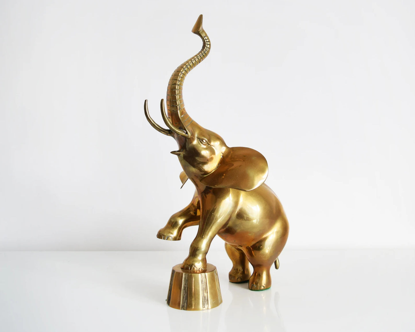 Another side front view of a large brass elephant that is balancing one of its front legs on a a platform and proudly displaying its trunk up for good luck.
